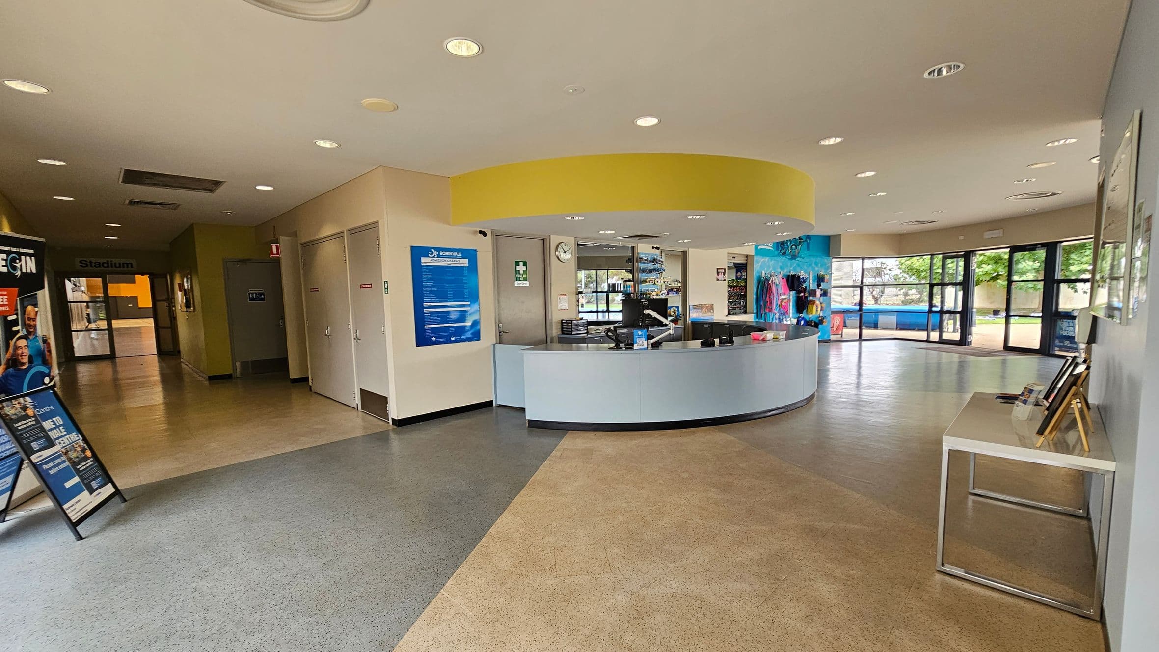 A spacious indoor area with a curved reception desk in the centre, featuring a computer and various items. To the left, a hallway leads to a door marked "Stadium," with posters and signs on the walls. On the right, large windows provide a view of greenery outside. The floor is a mix of green and beige tiles, and the ceiling is fitted with recessed lighting. A small table with brochures is positioned on the right side.