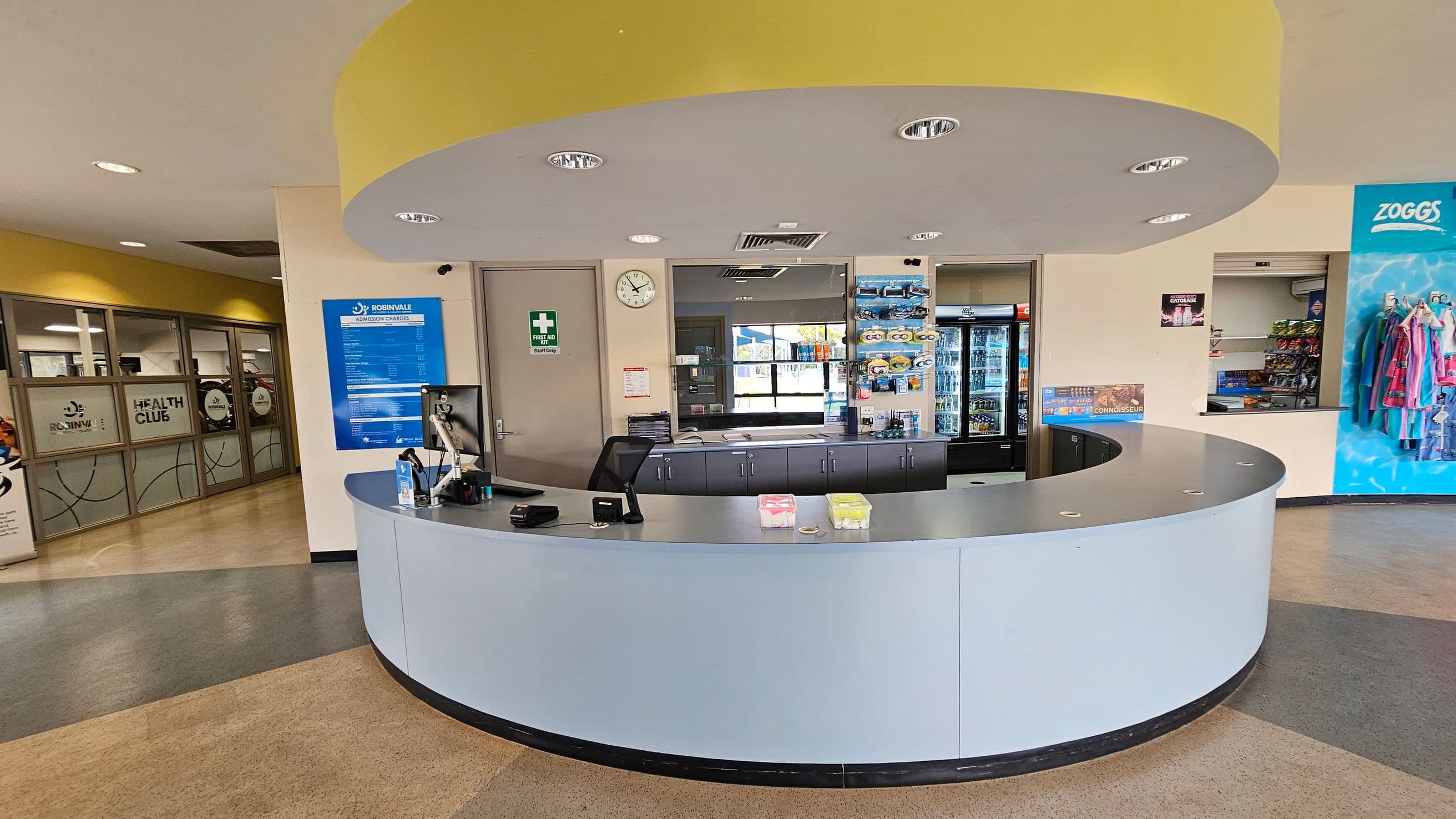 A reception area with a curved desk in the centre. Behind the desk, there's a wall with a clock, a first aid sign, and a door. To the right, a display of swimming accessories and a fridge with drinks. On the far right, a blue wall with hanging clothes. To the left, glass doors with "Health Club" signage. The ceiling is circular with recessed lights, and the floor has a mix of beige and green tiles.