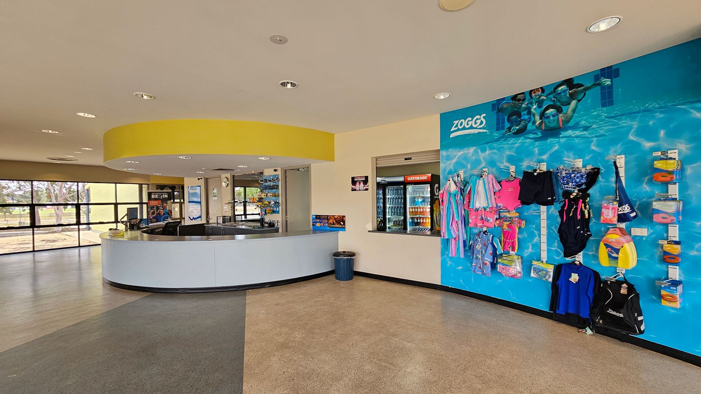 A reception area with a curved white desk on the left, topped with a yellow circular ceiling feature. Behind the desk, there are windows with a view of trees outside. On the right, a wall displays swimming gear, including swimsuits and accessories, against a blue background with an underwater image. A vending area with drinks is visible next to the display. The floor is a mix of grey and beige tiles.