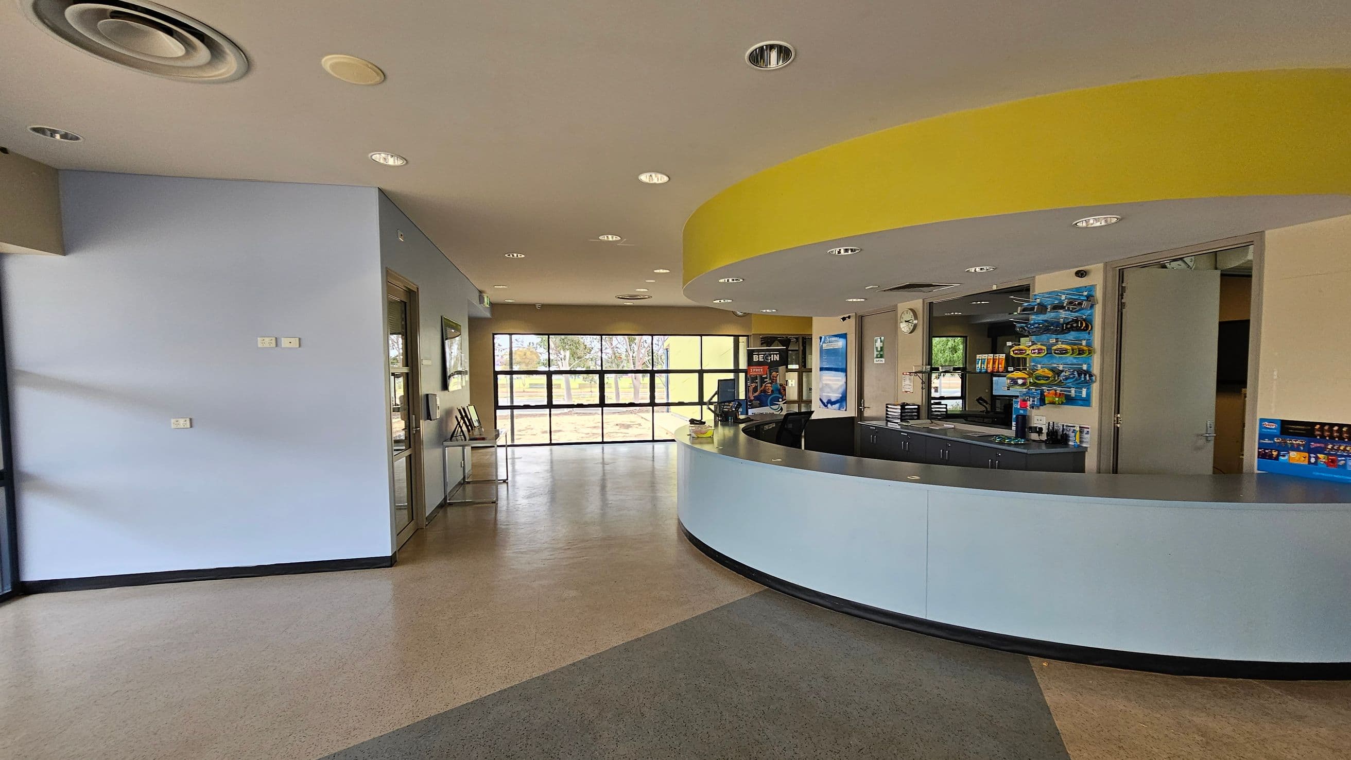 A spacious interior with a curved reception desk on the right, featuring a light blue front and a dark countertop. Behind the desk are shelves with various items. On the left, a light blue wall with a few electrical outlets. The floor is a mix of grey and green tiles. In the background, large windows provide a view of trees outside. The ceiling is beige with recessed lighting and circular vents.