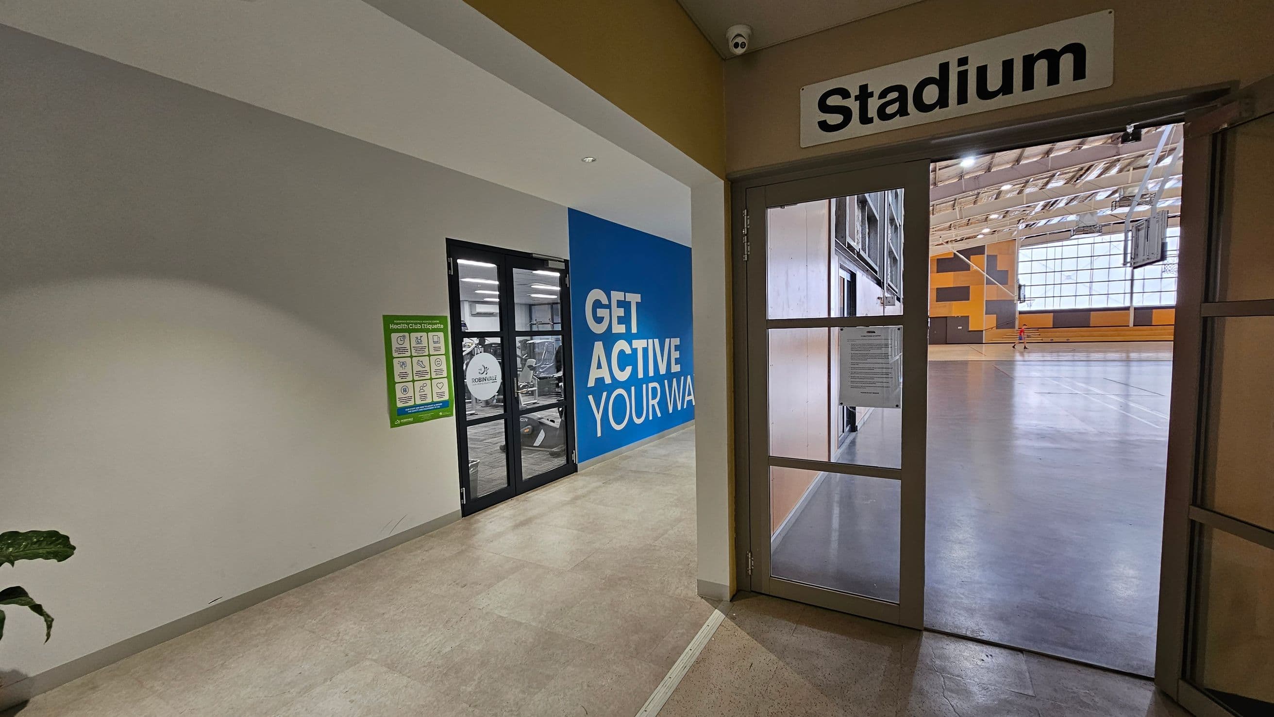 On the left, there's a hallway with a green wall and a poster about health club etiquette. A glass door leads to a gym area with exercise equipment. A large blue wall displays the words "GET ACTIVE YOUR WAY." On the right, a door marked "Stadium" opens into a spacious indoor sports area with a high ceiling, featuring a basketball court and large windows. The floor is a polished, light-coloured surface.