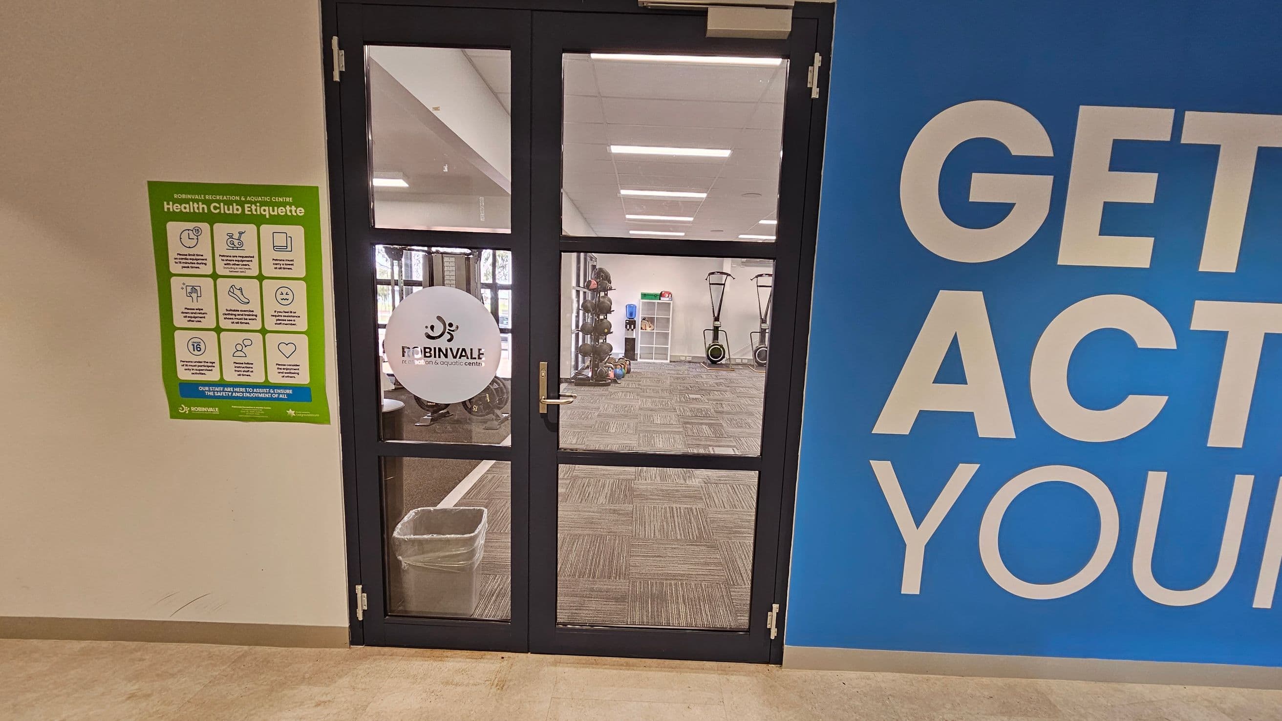 A glass door with a "ROBINVALE" logo leads to a gym area with exercise equipment, including weights and rowing machines. To the left of the door, a green poster outlines "Health Club Etiquette." On the right, a large blue wall displays partial text "GET ACT... YOUR." The floor is carpeted with a grey pattern, and the ceiling has rectangular lights.