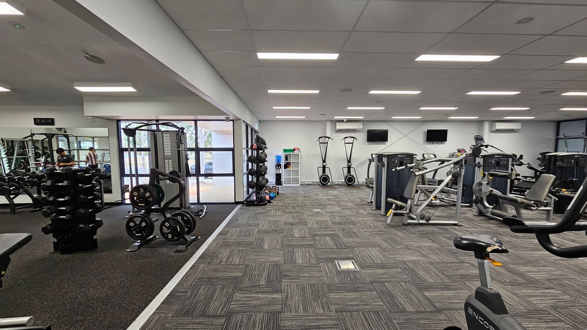 A gym interior with various exercise equipment. On the left, there are dumbbells and weight plates on racks, with a mirrored wall reflecting the equipment. In the centre, a window with a view outside and a shelf holding medicine balls and kettlebells. On the right, there are exercise machines, including stationary bikes and weight machines. The floor is covered with grey and green patterned carpet tiles, and the ceiling has rectangular fluorescent lights.