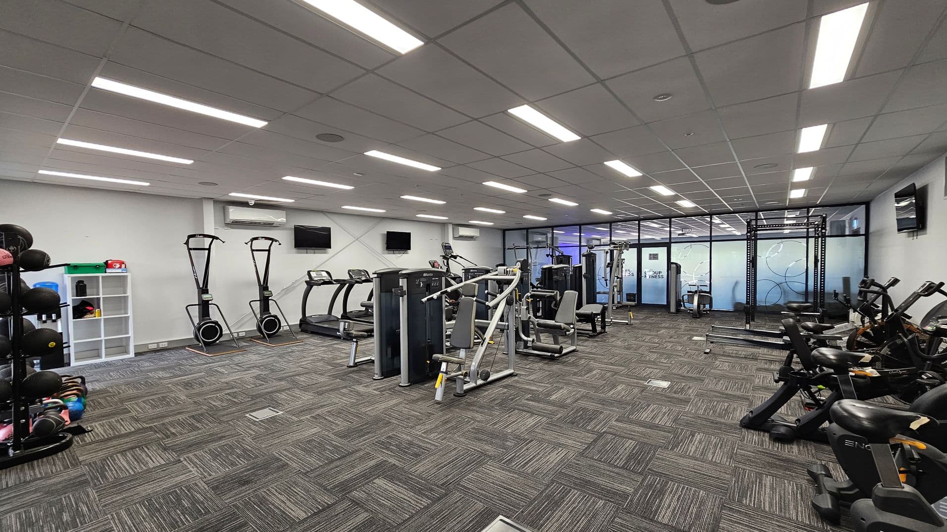 A modern gym with various exercise equipment. On the left, there are shelves with medicine balls and kettlebells. In the centre, there are treadmills and weight machines. On the right, stationary bikes are lined up. The floor has a grey and green patterned carpet, and the ceiling is fitted with rectangular fluorescent lights. The back wall features large glass panels with a frosted design.