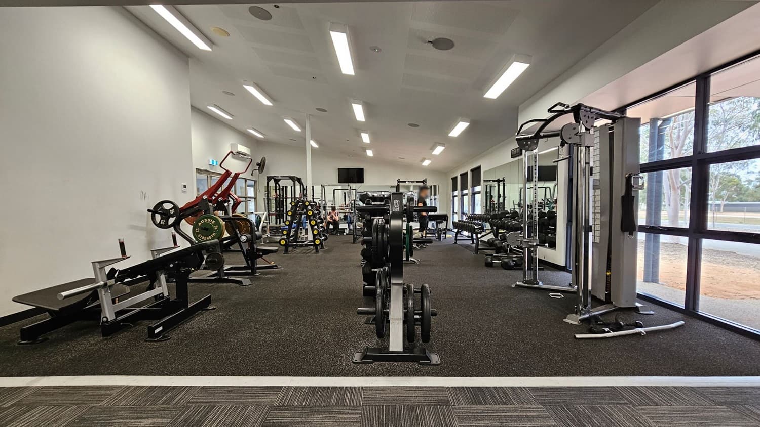 A gym with various exercise equipment. On the left, there are weight machines and benches. In the centre, a row of weight plates is visible. On the right, there is a cable machine near large windows that let in natural light. The floor is covered with dark rubber mats, and the ceiling has bright fluorescent lights.