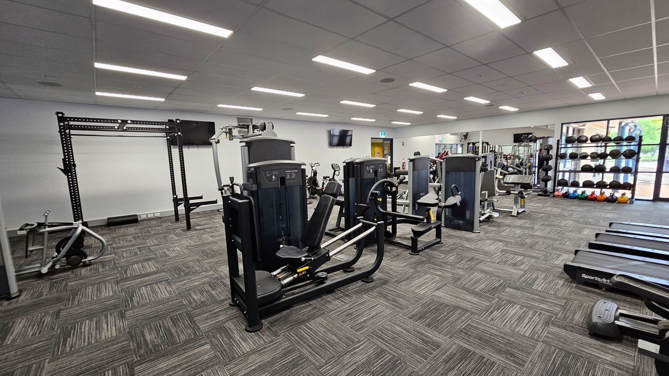 A gym with various exercise equipment. On the left, there's a squat rack and a rowing machine. In the centre, several weight machines are arranged. On the right, treadmills are lined up, and a rack of medicine balls and kettlebells is visible near large windows. The floor is covered with patterned carpet tiles, and the ceiling has recessed lighting.
