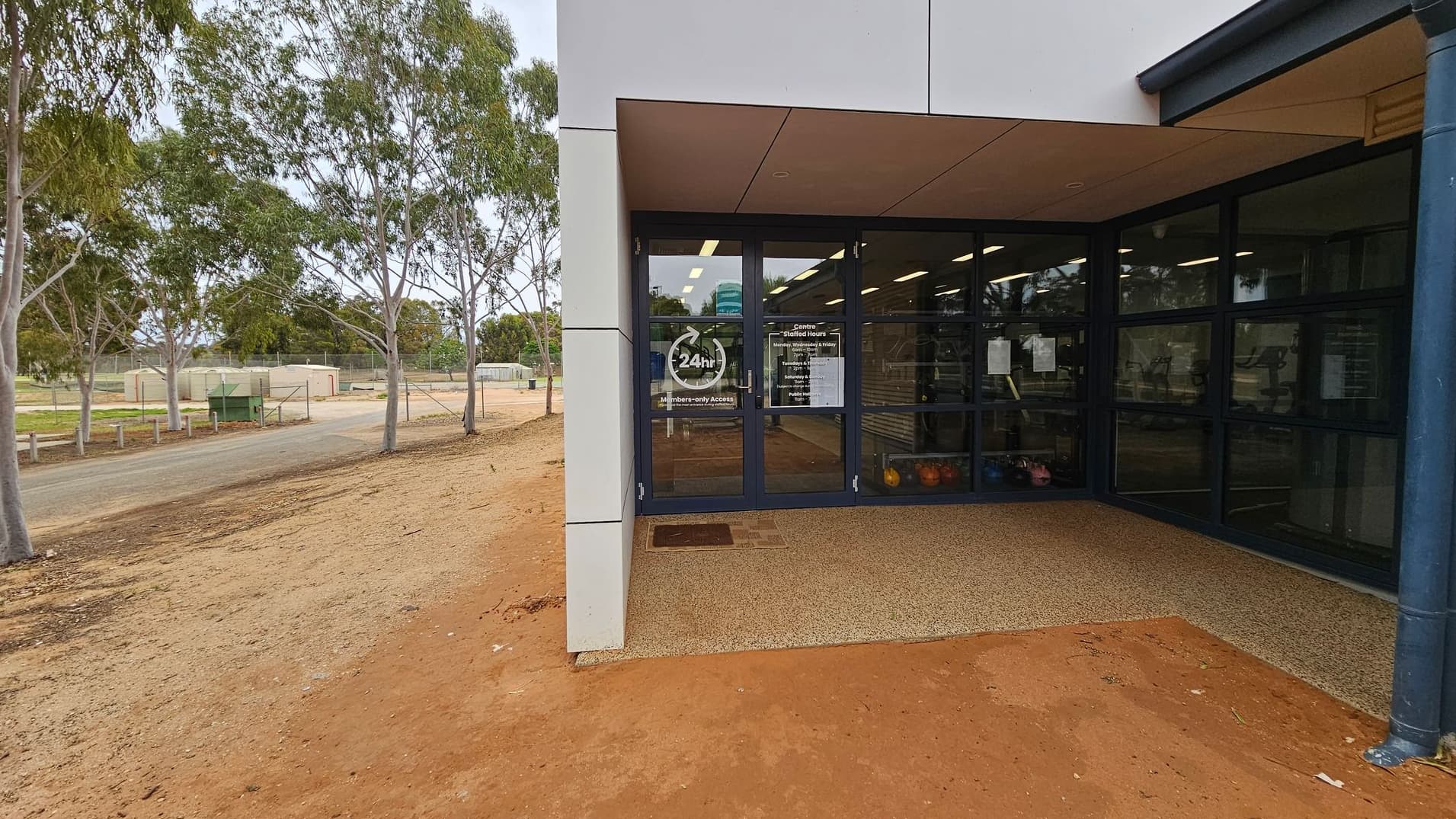 A building entrance with glass doors and windows on the left, displaying signs and reflections. The ground is paved with tiles. On the right, there is a dirt path with sparse grass and several trees. In the background, a structure with a green roof is visible. The ceiling above the entrance is textured.