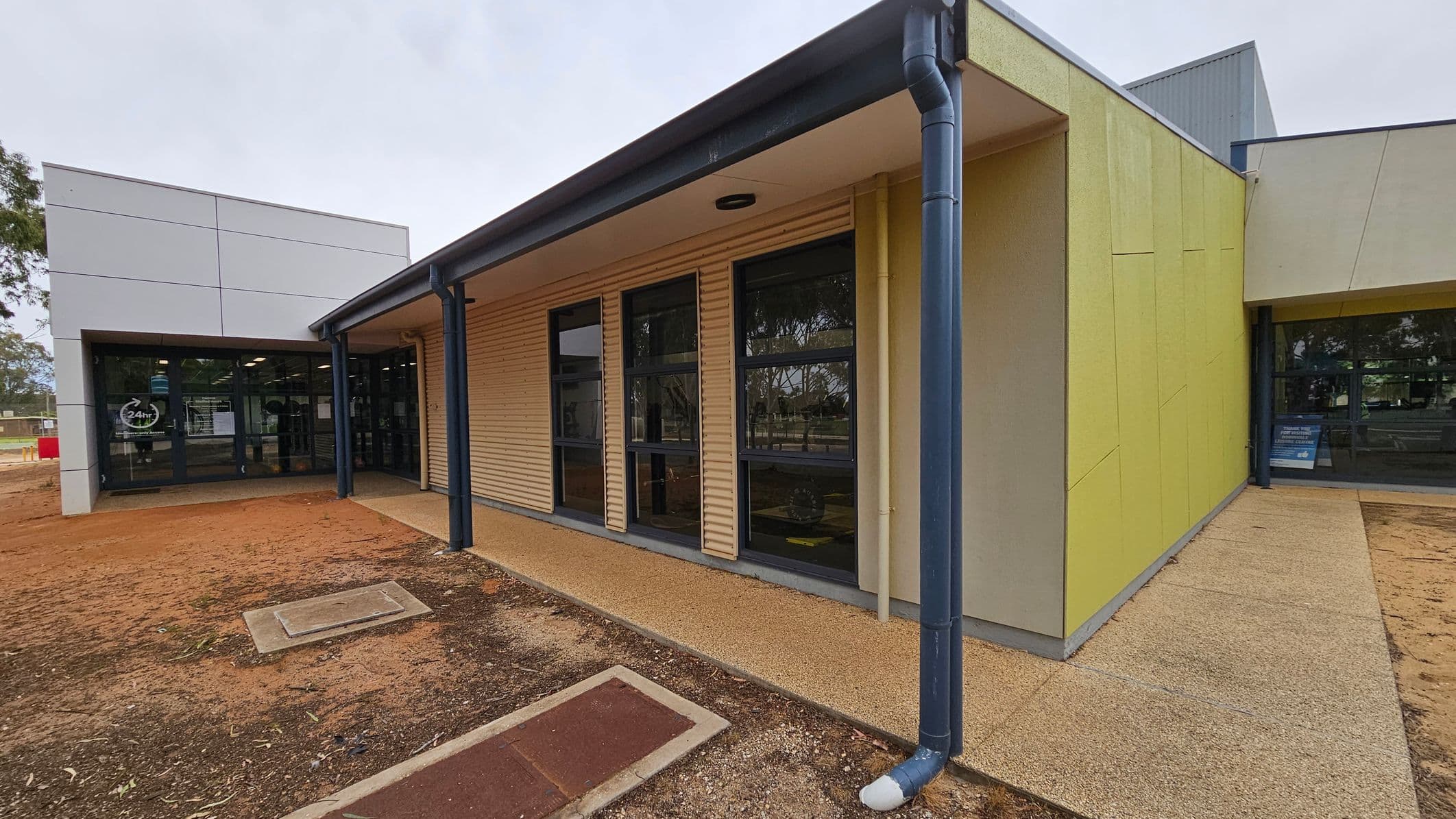 A modern building with a combination of beige and light green walls. On the left, there is a section with large glass windows and a door, displaying a "24hr" sign. The middle section features corrugated beige panels with three tall windows. The right side has a light green wall with a downpipe. The ground is a mix of concrete and reddish-brown soil, with a rectangular metal cover. Trees are visible in the background through the windows.