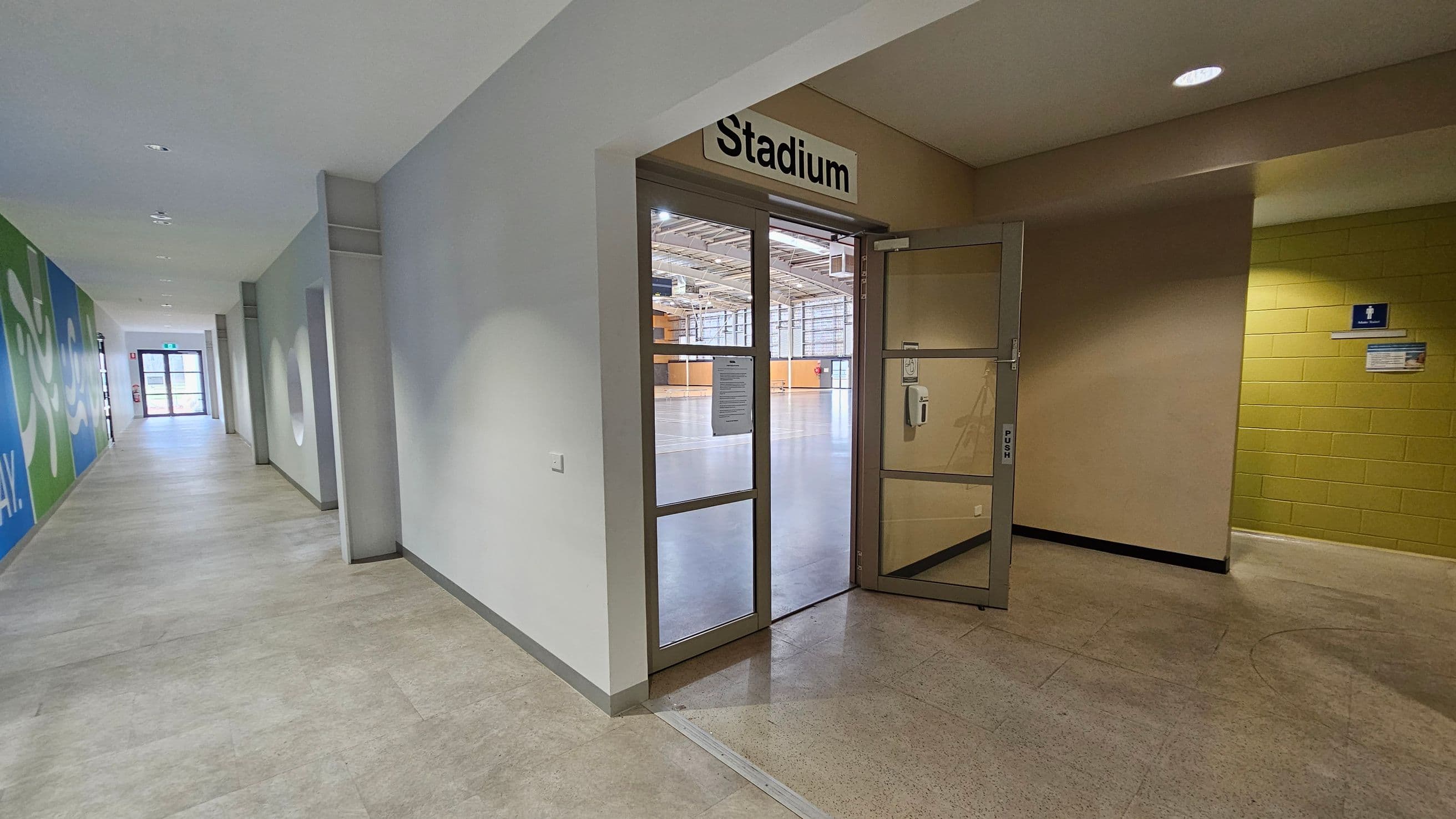 A hallway with a door labelled "Stadium" leading to a large indoor sports area. The door is partially open, revealing a spacious gymnasium with a high ceiling and geometric wall patterns. On the left, there's a glass wall with a view into a fitness centre, featuring exercise equipment and a large blue sign that reads "GET ACTIVE YOUR WAY". The floor is tiled, and a hand sanitiser dispenser is mounted on the wall near the door.