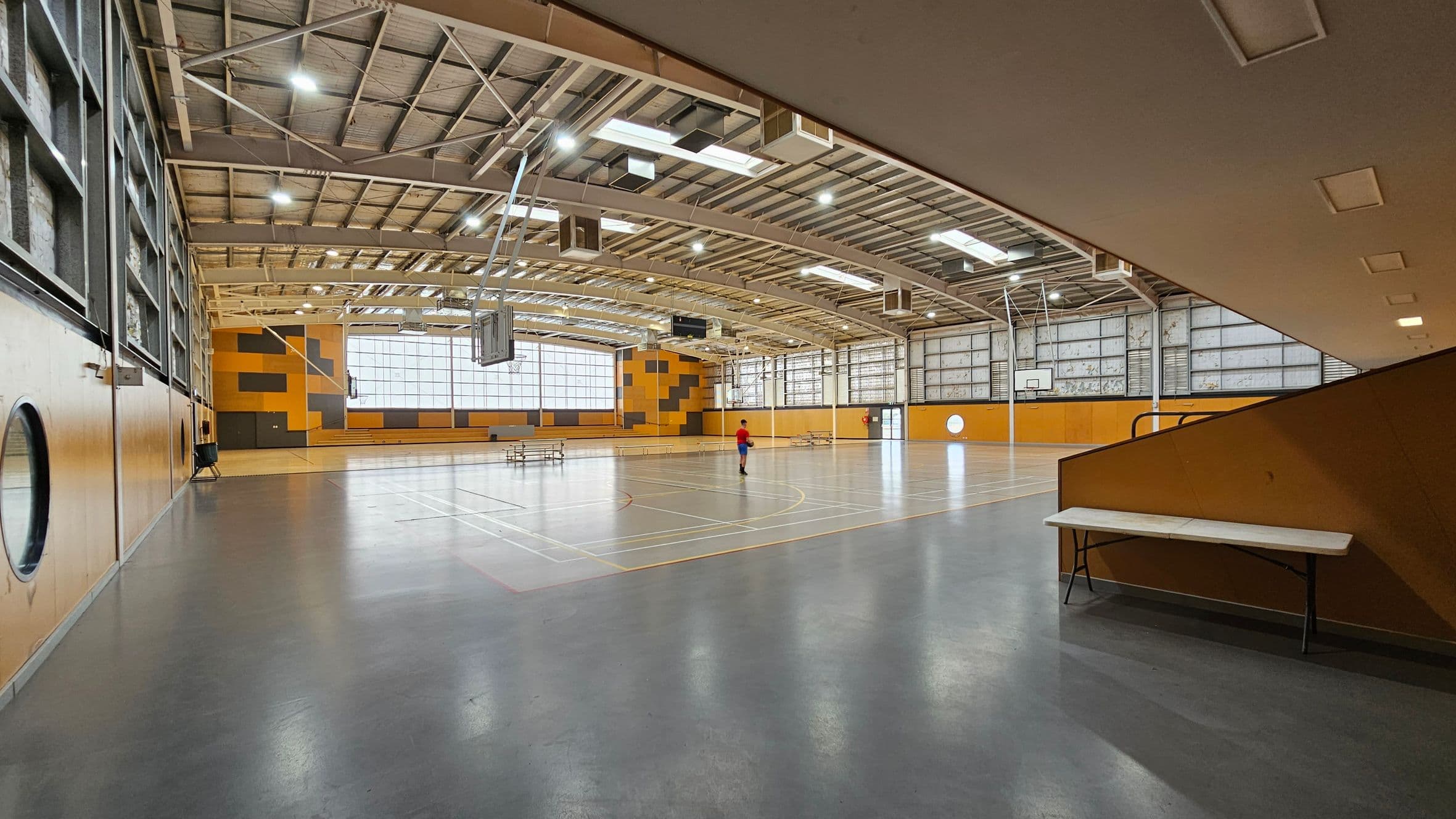 A spacious indoor sports hall with a high, metal-beamed ceiling and large windows on the far wall. The floor is marked for various sports. A single person in a red shirt stands in the middle. On the right, a white folding table is against a brown wall. The walls are a mix of yellow and grey panels. Basketball hoops are visible, and the lighting is bright.