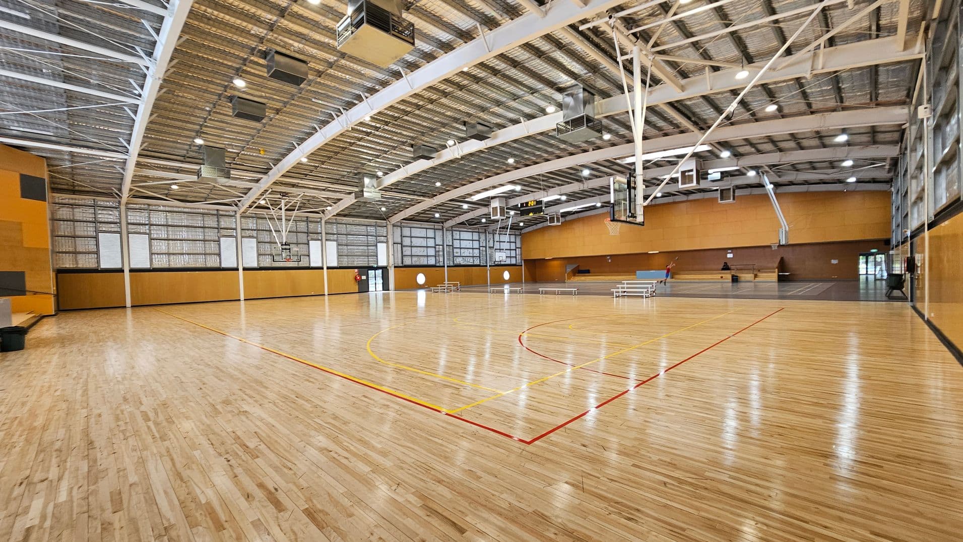 A spacious indoor basketball court with a polished wooden floor marked with red and yellow lines. The ceiling is high with exposed metal beams and lighting fixtures. On the right, there are basketball hoops and a scoreboard. The left side features large windows with metal frames. In the background, there are bleachers and a small entrance. The walls are painted in a warm yellow-brown colour.
