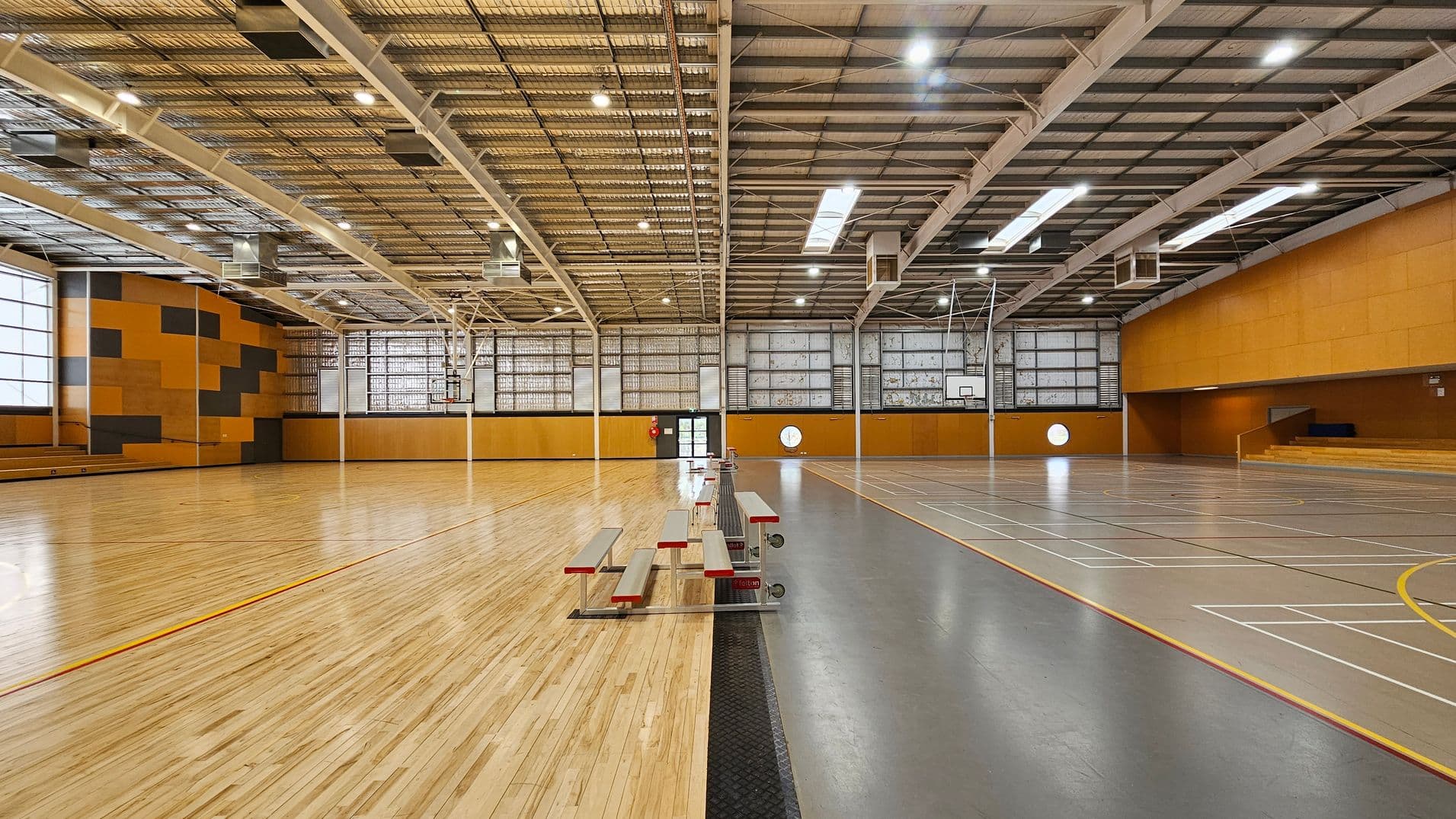A spacious indoor sports hall with a wooden floor on the left and a grey sports court on the right. The ceiling is high with exposed metal beams and bright lighting. The walls are a mix of orange and grey panels, with large windows allowing natural light. In the centre, there are retractable bleachers. A basketball hoop is visible on the right side. The hall is empty, providing a clear view of the entire space.