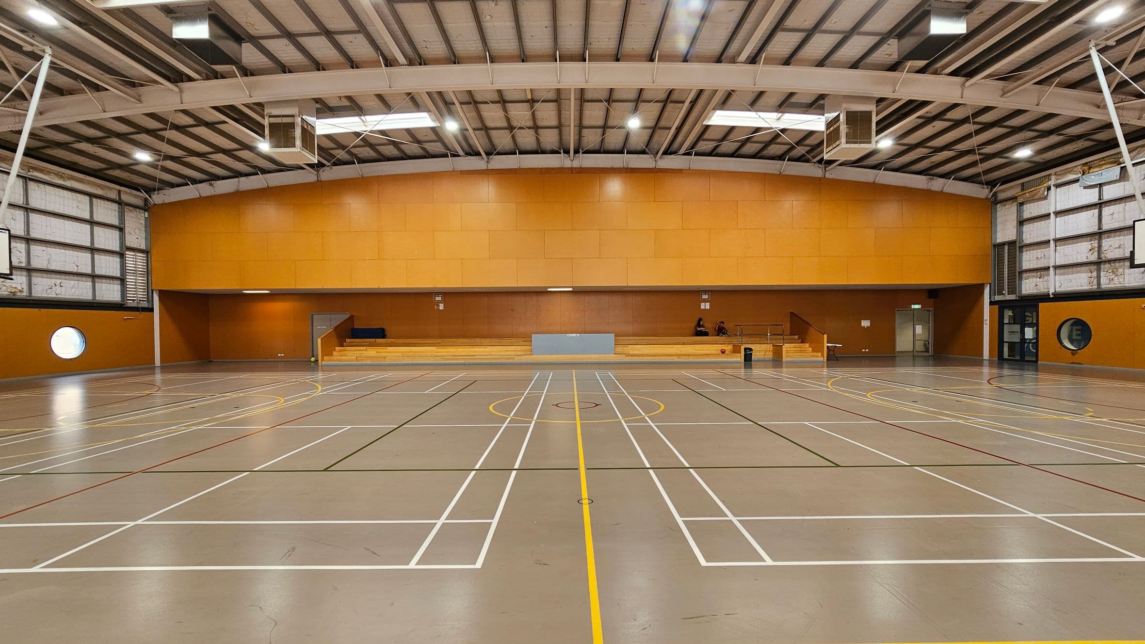 A spacious indoor sports hall with a grey floor marked with various sports lines in white, yellow, and red. The walls are orange, and there are large windows on the left and right. In the centre, there are wooden bleachers with a few people seated. The ceiling is high with exposed beams and lighting fixtures. A basketball hoop is visible on the left wall, and there are circular windows on both sides.