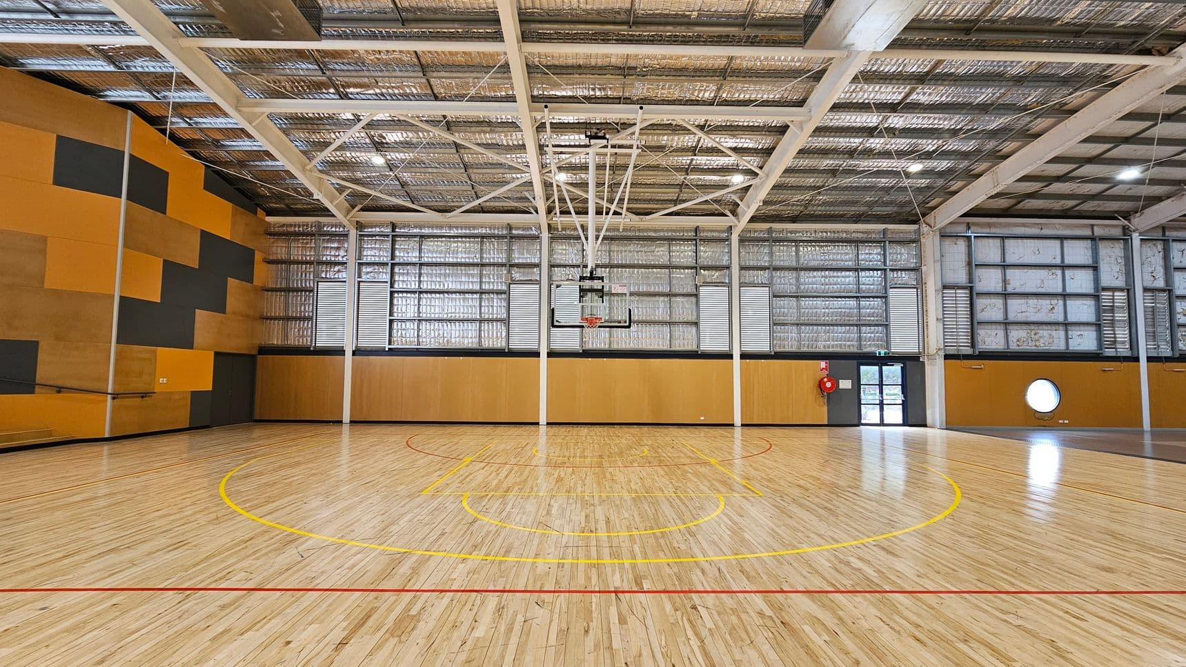 A spacious indoor basketball court with a polished wooden floor marked with red and yellow lines. In the centre, a basketball hoop is suspended from the ceiling. The left wall features a geometric pattern in orange and dark grey. The back wall has large windows with metal frames, and a door with a fire extinguisher nearby. The ceiling is high with exposed metal beams and insulation.