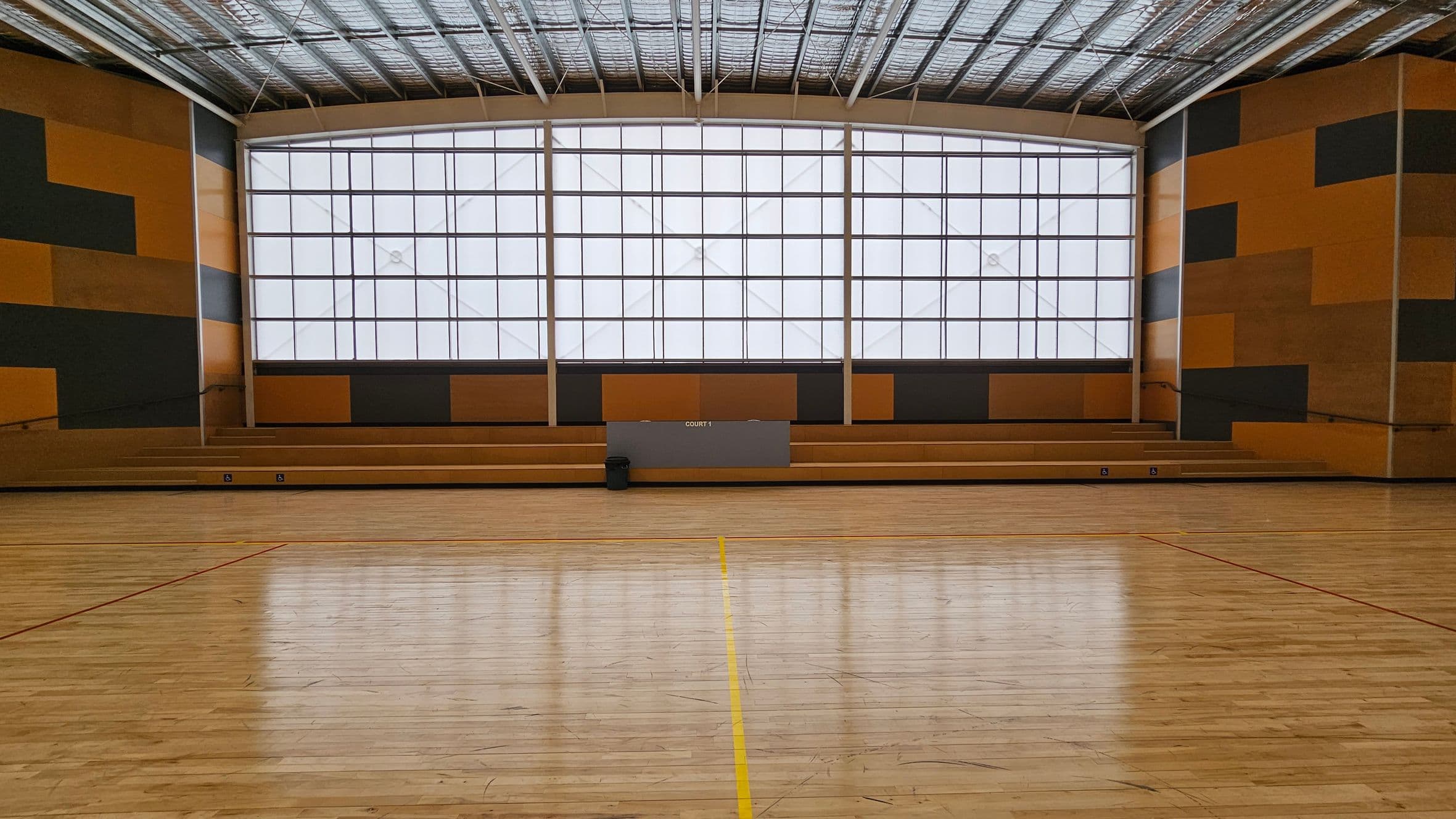 A spacious indoor sports hall with a polished wooden floor marked with red and yellow lines. The back wall features large, grid-patterned windows allowing diffused light. The walls are decorated with a geometric pattern of orange and dark grey panels. Wooden bleachers are positioned against the back wall, with a small bin placed on the floor. The ceiling is high with visible metal beams and insulation.