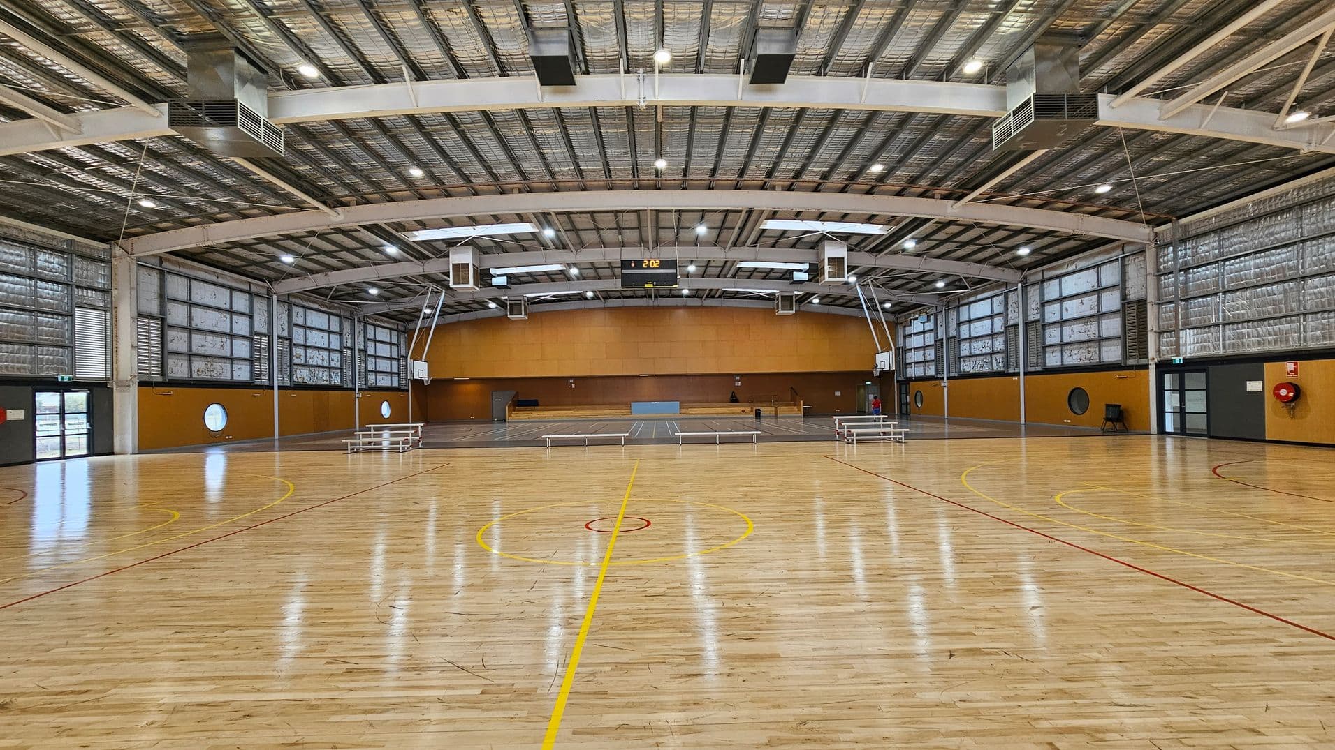 A spacious indoor sports hall with a polished wooden floor marked for basketball. The ceiling is high with exposed metal beams and lighting fixtures. On the left and right, there are large windows with metal grilles. Two basketball hoops are visible, one on each side. The back wall is orange with a digital clock displaying "2:02" at the centre. Benches are placed along the sides, and there are doors on both ends of the hall.