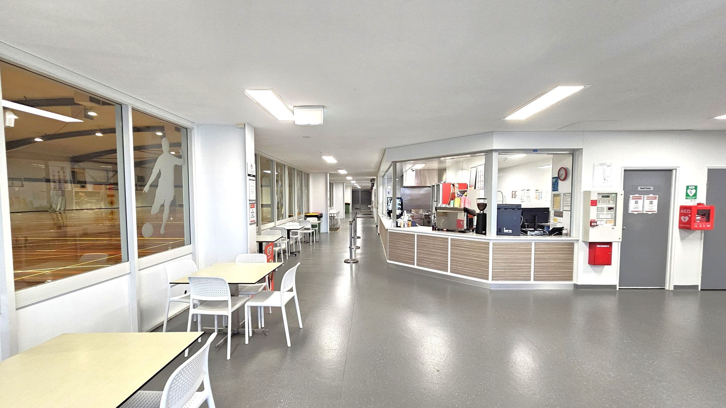 A spacious indoor area with a grey floor and white ceiling. On the left, large windows reveal a sports hall with a silhouette of a footballer. Below the windows, there are tables and white chairs. On the right, a counter with kitchen equipment and a coffee machine is visible, along with a grey door marked "Staff Only". A red AED box is mounted on the wall. The area is well-lit with ceiling lights, and a barrier guides foot traffic near the counter.