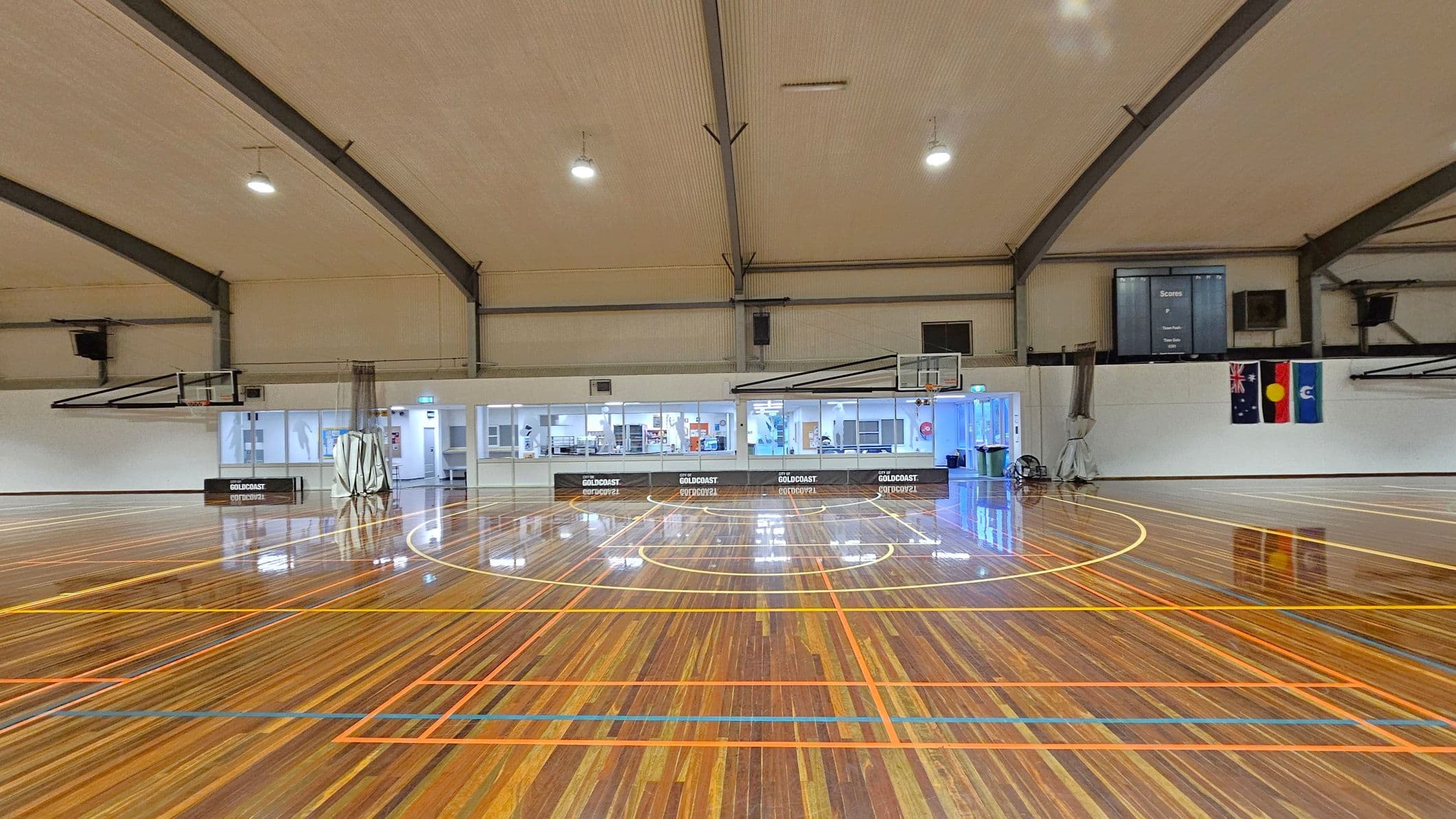 A spacious indoor basketball court with a polished wooden floor featuring orange and yellow lines. Two basketball hoops are visible on either side. The ceiling is high with exposed beams and overhead lights. The back wall has a row of windows and doors, with a scoreboard and three flags displayed. The area is well-lit, and the reflections on the floor are prominent.