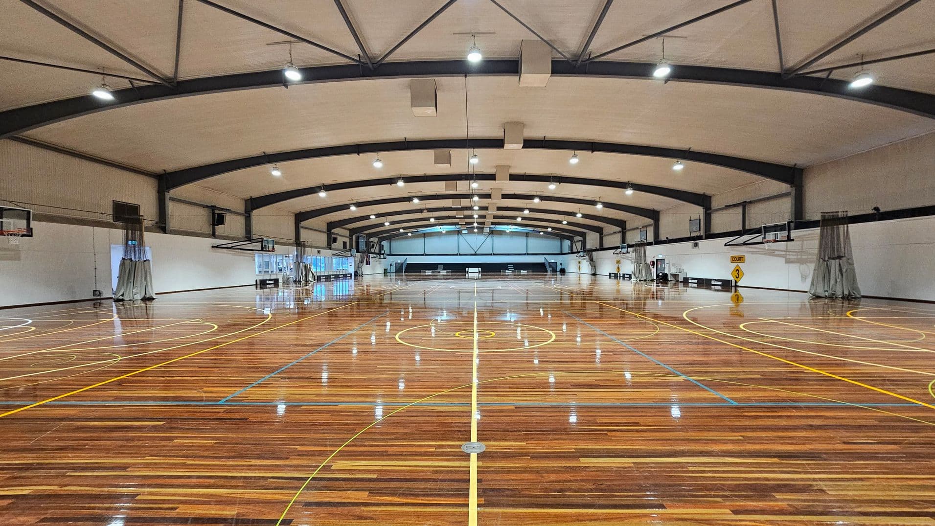 A spacious indoor basketball court with a polished wooden floor featuring yellow and blue court markings. The ceiling is high with exposed beams and multiple overhead lights. On both sides, there are basketball hoops and white walls with some folded curtains. The far end of the court has a set of windows and a scoreboard. A sign indicating "Court 3" is visible on the right side.