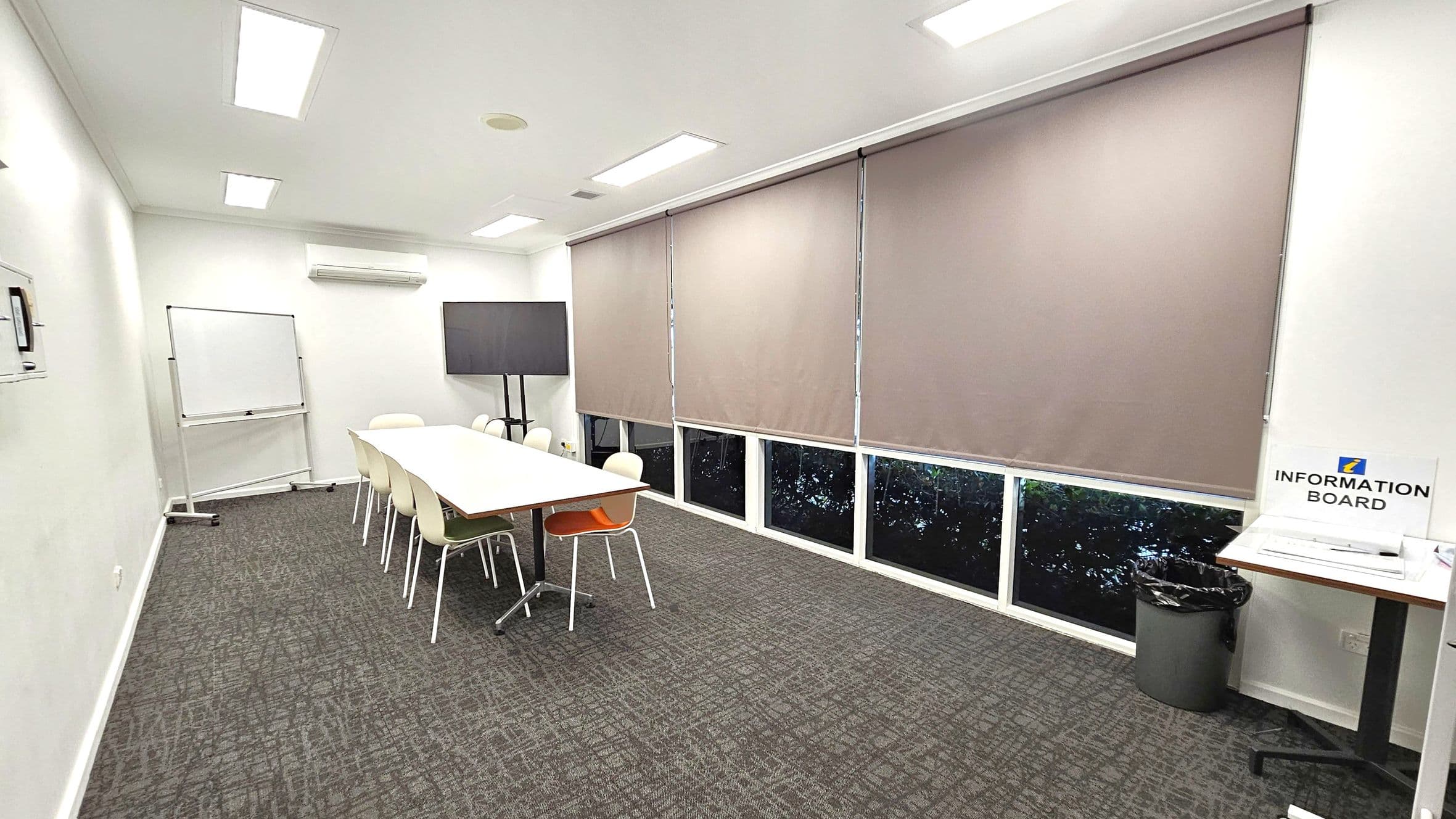 A conference room with a long white table surrounded by chairs, including one orange chair. On the left, there's a whiteboard on wheels. The right side features large windows covered with beige blinds. A flat-screen TV is mounted on a stand near the corner. An "Information Board" sign is on a small table with a bin underneath. The floor is carpeted in a grey pattern, and the ceiling has recessed lighting.