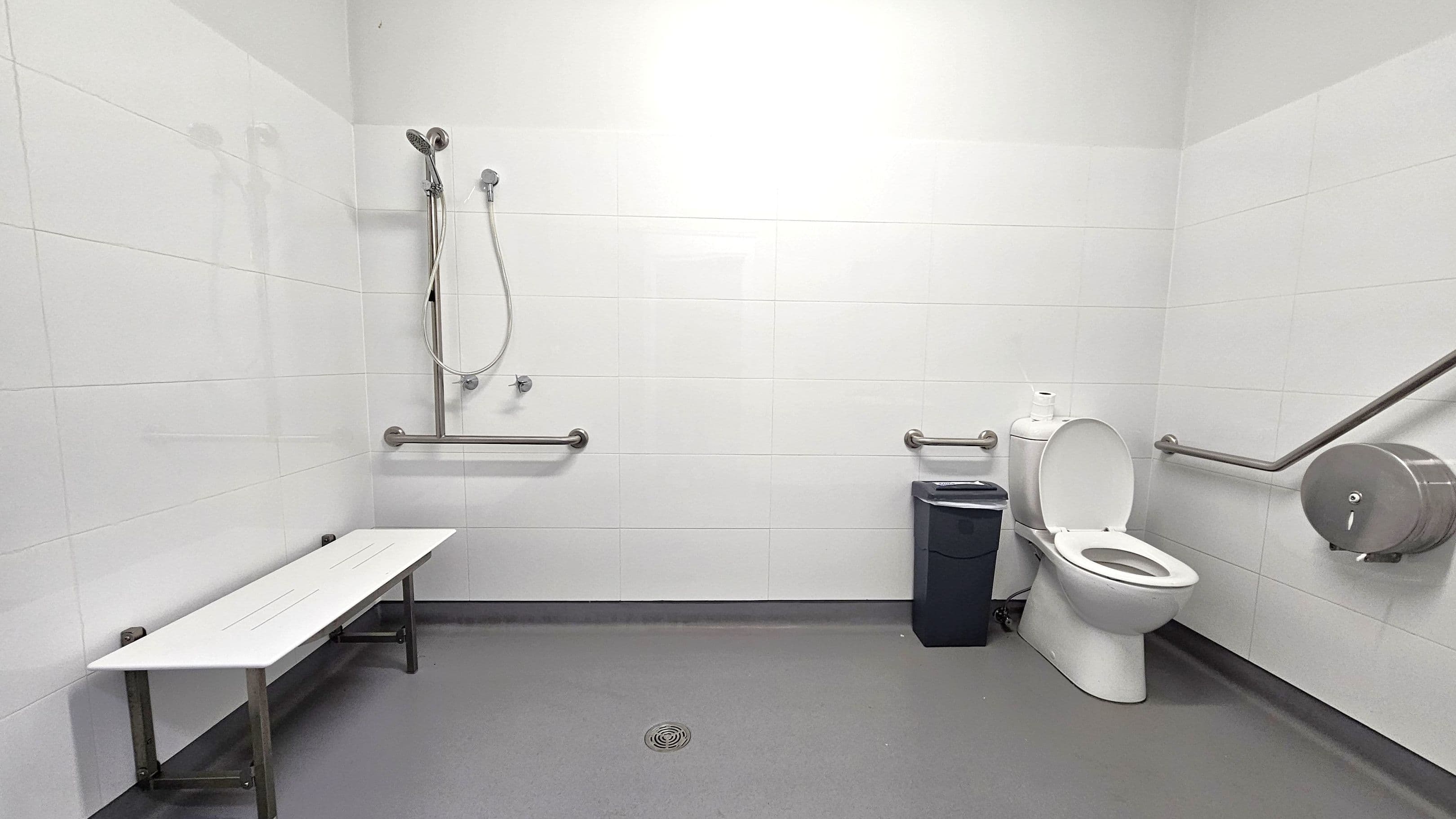 A spacious, accessible bathroom with white tiled walls and a grey floor. On the left, a white bench is attached to the wall. In the centre, a showerhead with a flexible hose is mounted on the wall, accompanied by horizontal grab bars. On the right, a toilet with an open lid is next to a black bin. A large toilet paper dispenser and additional grab bars are mounted on the wall near the toilet. The ceiling is plain and white.