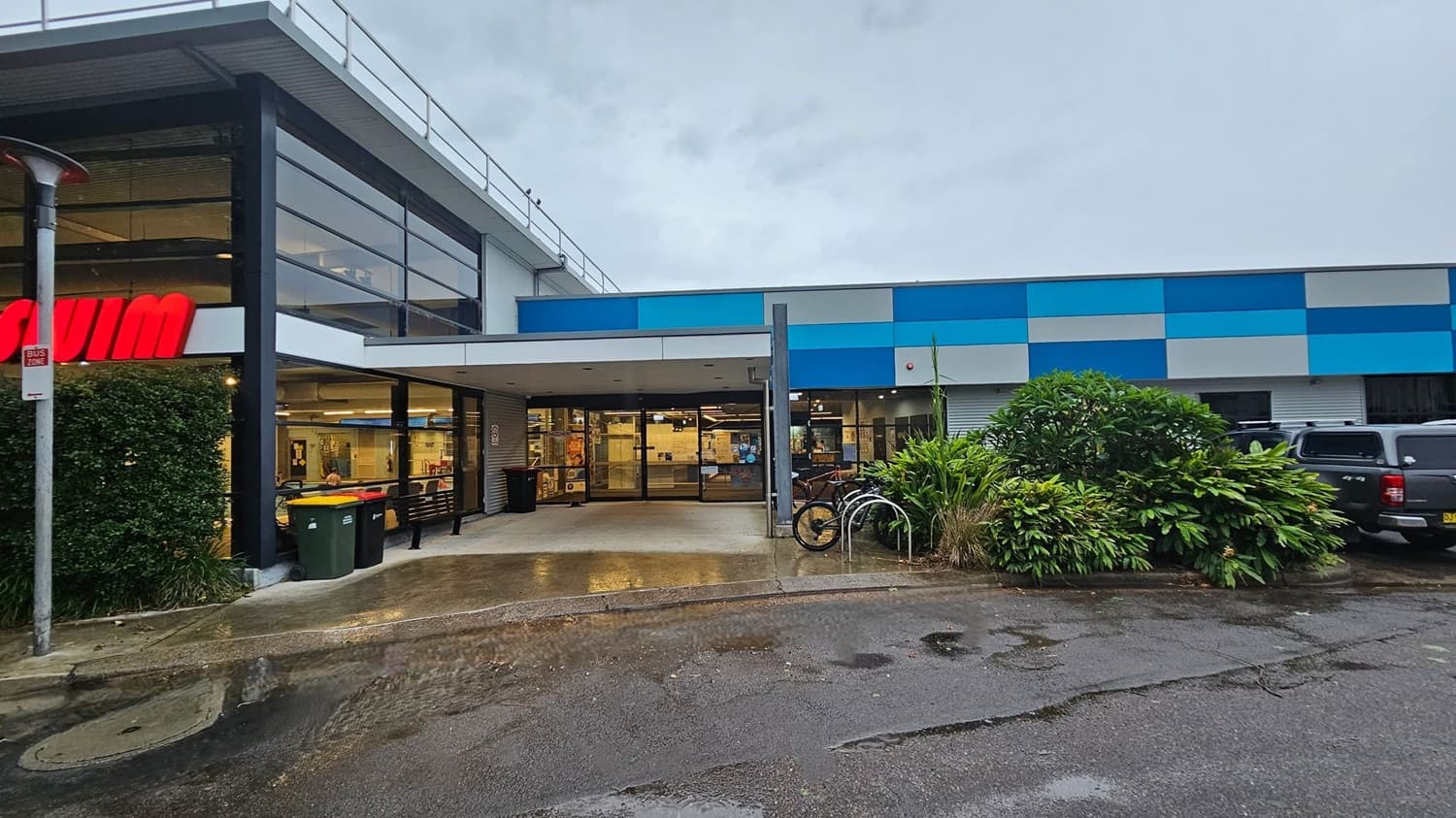 A car park with several vehicles is on the left, bordered by a grassy area and trees. On the right, a building with a blue and grey facade has large windows and a few parked cars in front. The ground is wet with puddles, and there are yellow bollards near the building. In the background, more trees and a grassy field are visible.