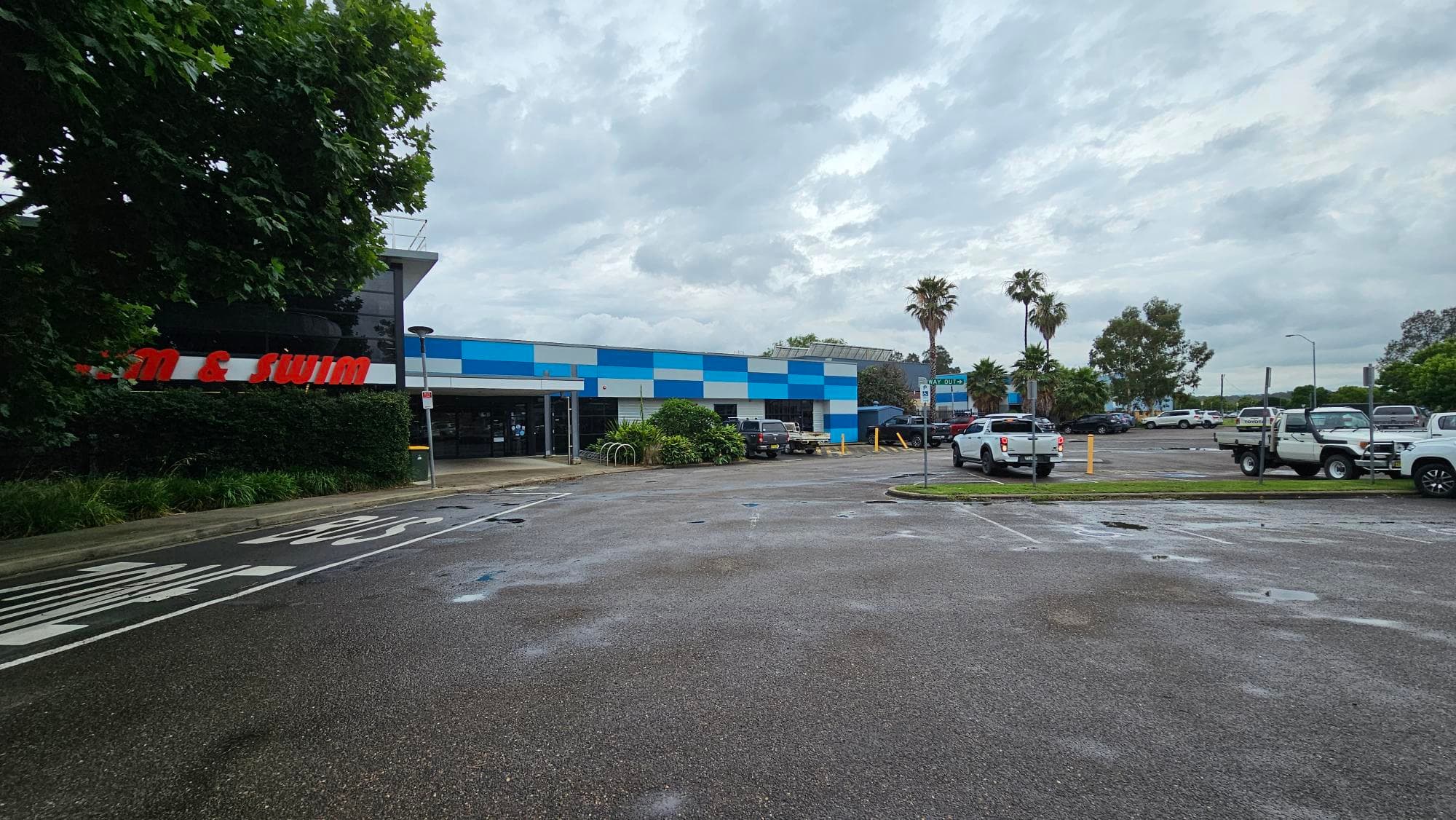 A car park with several vehicles, including white trucks, is in the foreground. On the left, a building with a partially visible red sign is surrounded by greenery. The building has a blue and white checkered pattern. In the background, there are palm trees and more parked cars. The sky is overcast.