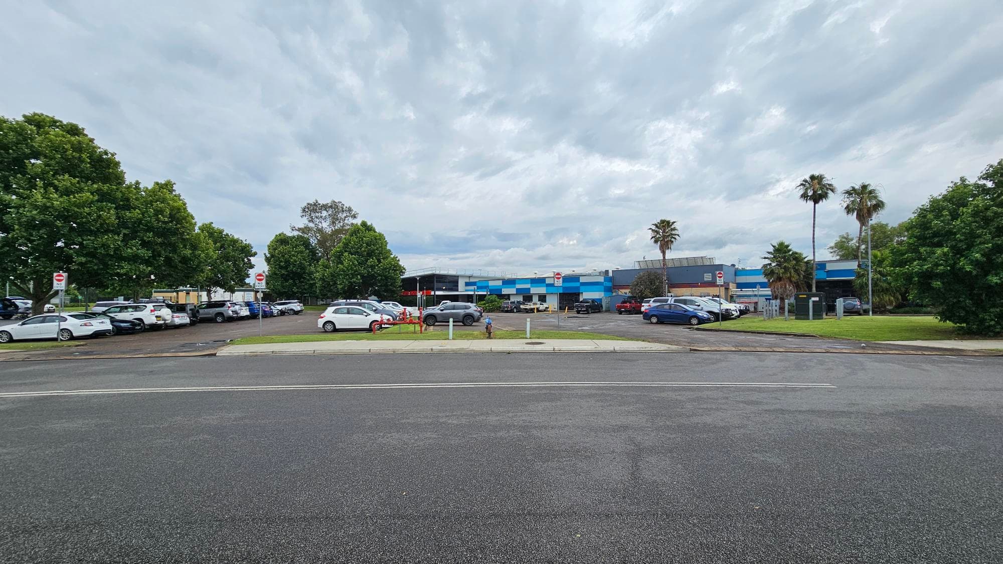 A car park with several vehicles is in the foreground, bordered by green grass and trees on both sides. In the background, there is a blue and grey industrial building. The sky is overcast, and there are palm trees near the building. The road in the foreground is empty, with a clear view of the parking area and the building beyond.