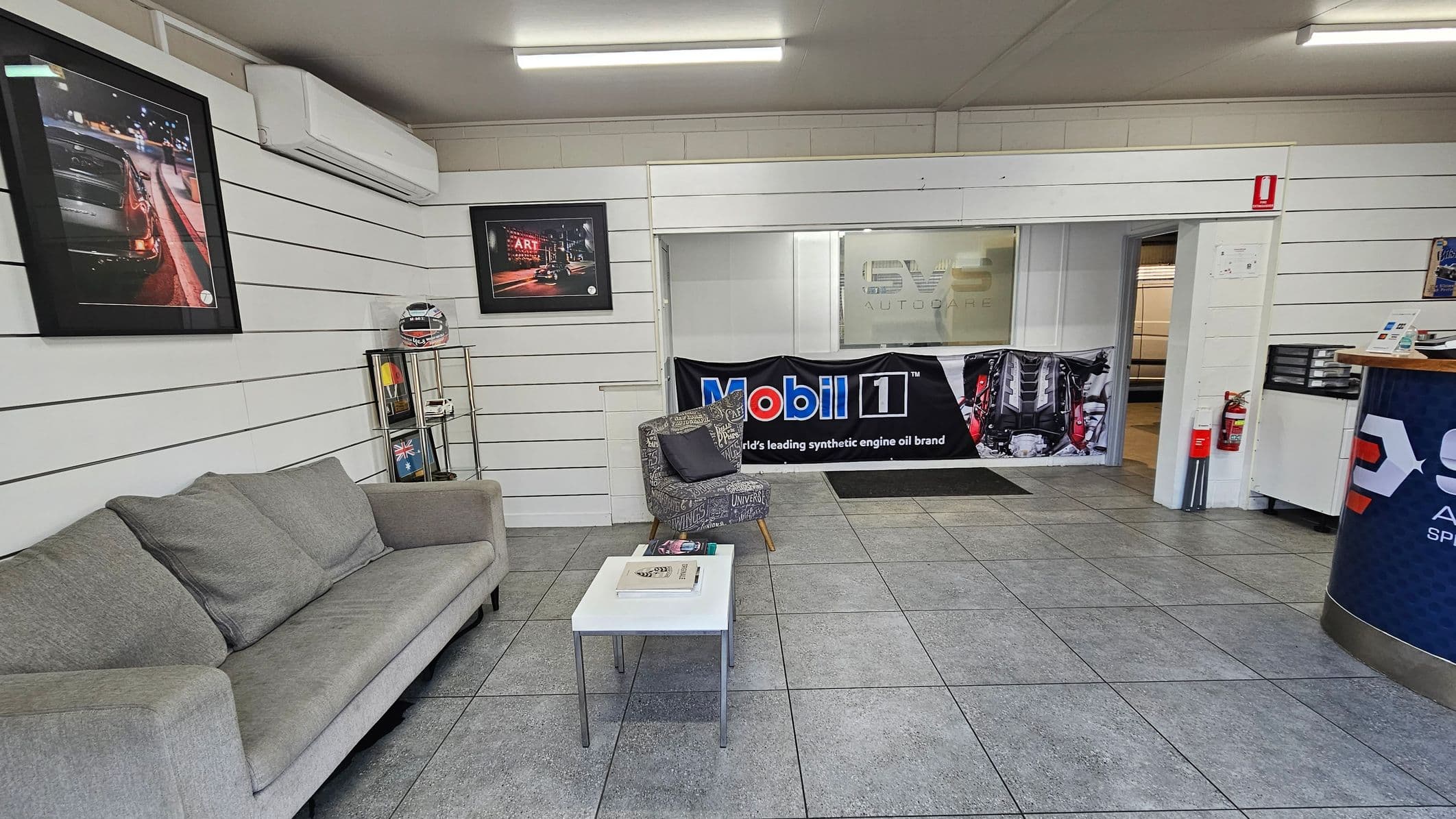 A building with a sign reading "SVS Autocare Specialist Vehicle Servicing" above the entrance. The office door is in the centre, with a red "OFFICE" sign above it. A potted plant is to the left of the door, and two green bollards are in front. A window is on the right side of the entrance. A truck is partially visible on the left, near a roller door with a "PARKING" sign. The ground is paved with some yellow markings.