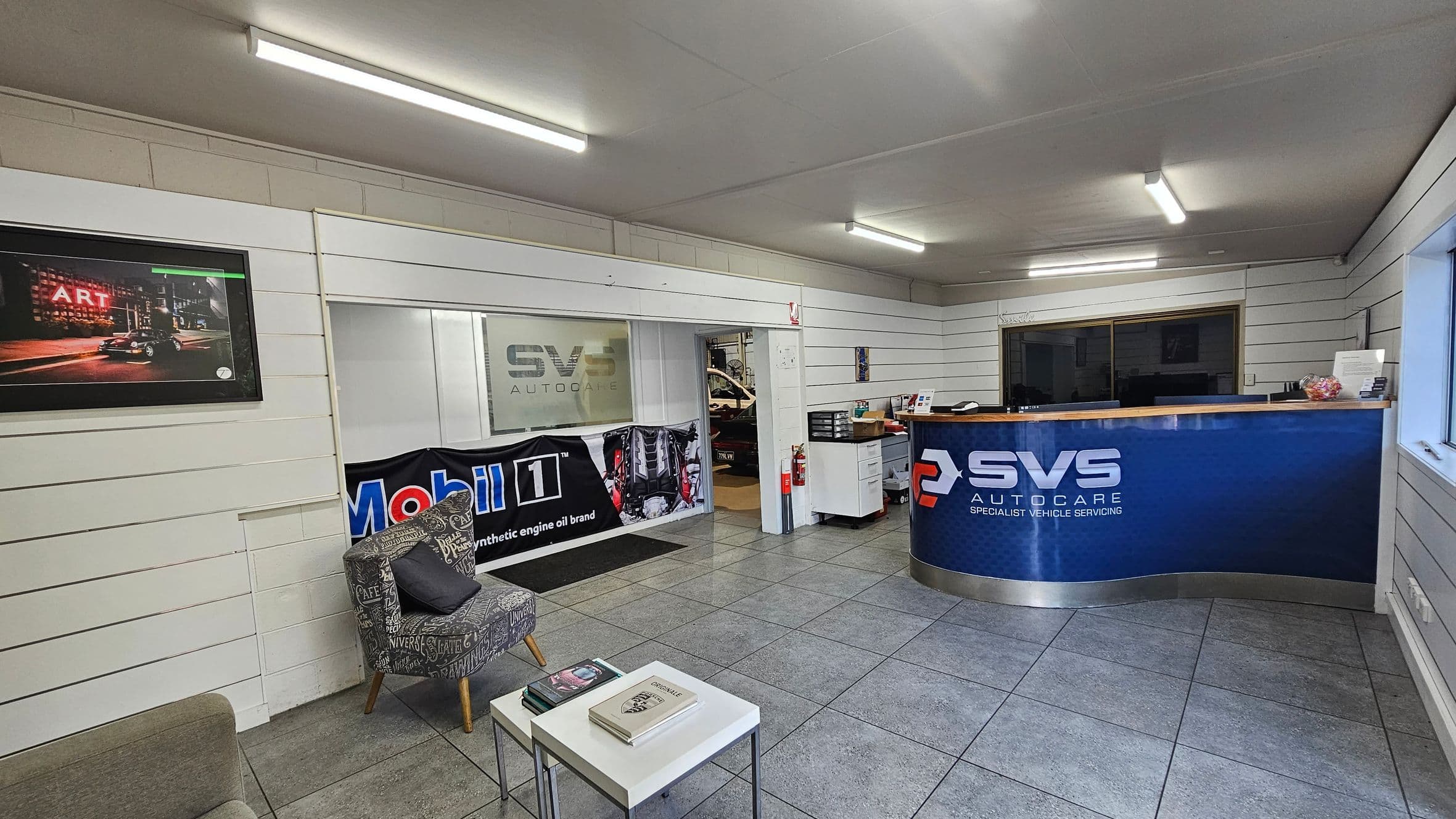 A modern automotive service reception area with grey tiled flooring and a white panelled ceiling. On the left, there's a wall-mounted TV above a patterned armchair and a small white table with magazines. A banner for Mobil 1 is displayed near a window with "SVS Autocare" signage. On the right, a blue reception desk with the SVS Autocare logo is positioned in front of a large window. The room is well-lit with fluorescent lights.