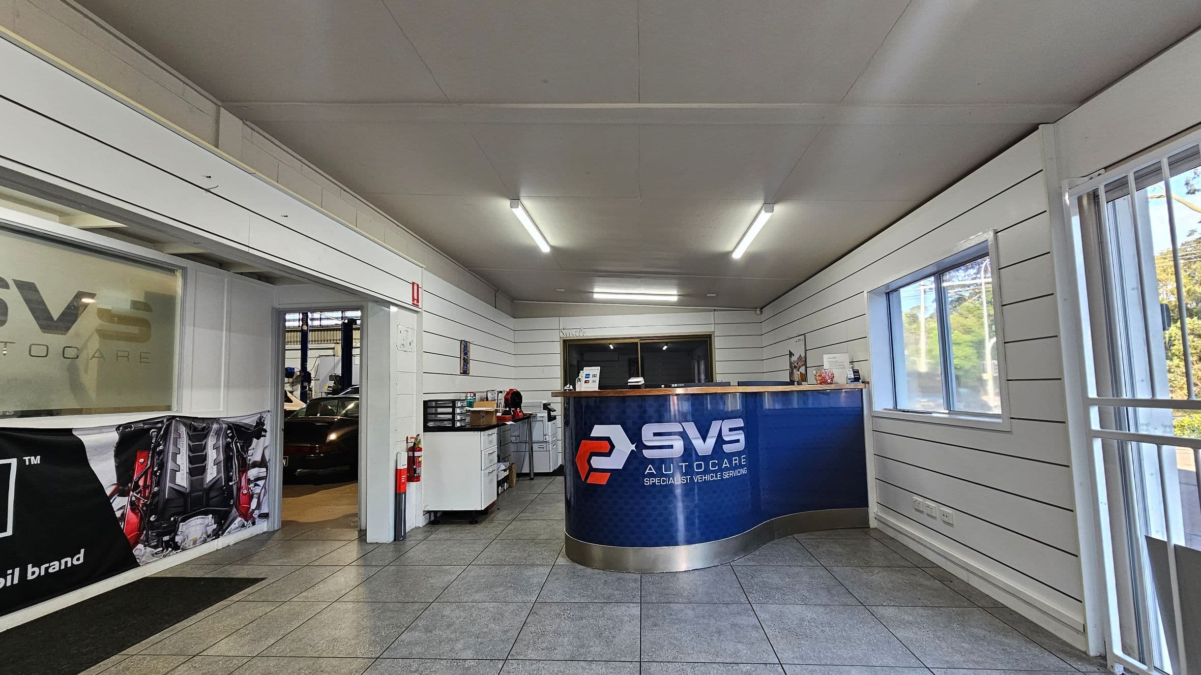 A reception area with a blue and silver curved counter on the right, displaying the logo "SVS Autocare". The walls are white with horizontal panels. On the left, there's a doorway leading to a garage with a car visible. A banner with an engine image is hung near the door. A small desk with office supplies and a fire extinguisher is next to the door. The floor is tiled, and the ceiling has fluorescent lights. A window on the right wall shows trees outside.