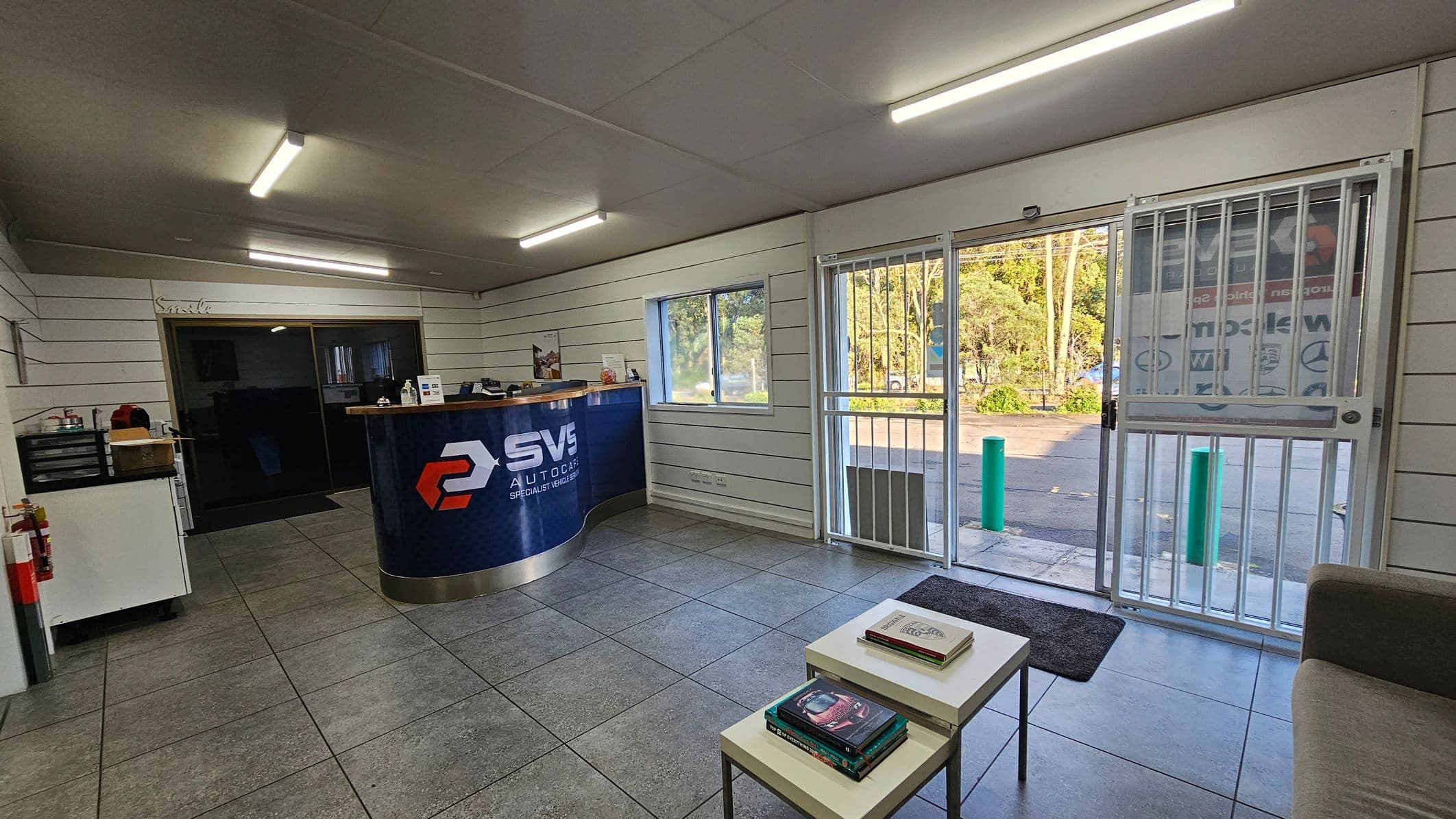 A reception area with a tiled floor and fluorescent lighting on the ceiling. On the left, there's a counter with a logo and various items on top. Behind it, a small office area is visible. On the right, a glass door with metal bars leads outside, where trees and a parking area can be seen. In the foreground, a small table with books is next to a grey sofa. The walls are lined with horizontal panels, and a window is present near the counter.