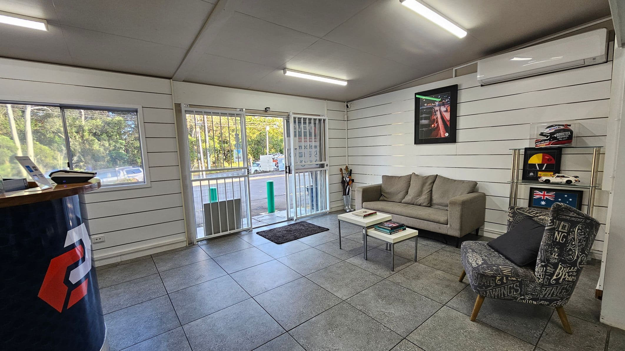 A room with grey tiled flooring and a white panelled ceiling. On the left, there's a counter with a device on top. The back wall has a large window and a glass door with bars, leading outside. On the right, a beige sofa sits against the wall, with a small table in front holding books. A patterned armchair is nearby. Above the sofa, a framed picture hangs, and a shelf displays a helmet, model cars, and an Australian flag. An air conditioning unit is mounted above.