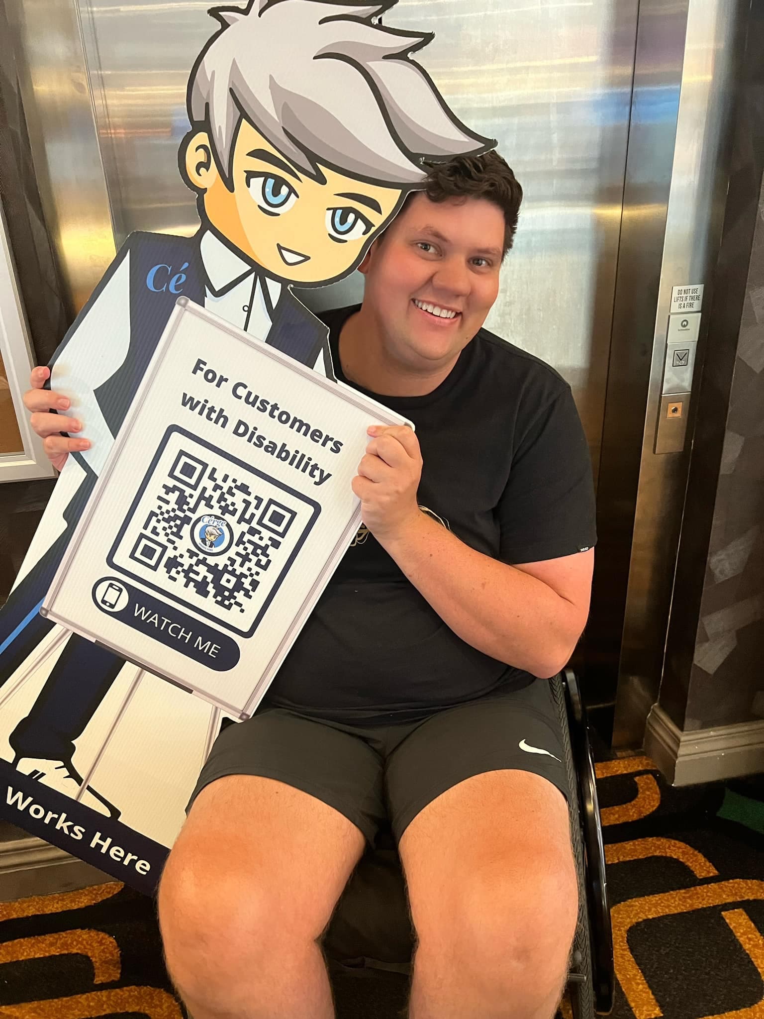 A man in a suit sits in a mobility chair beside a Cérge cardboard cutout with a QR sign.