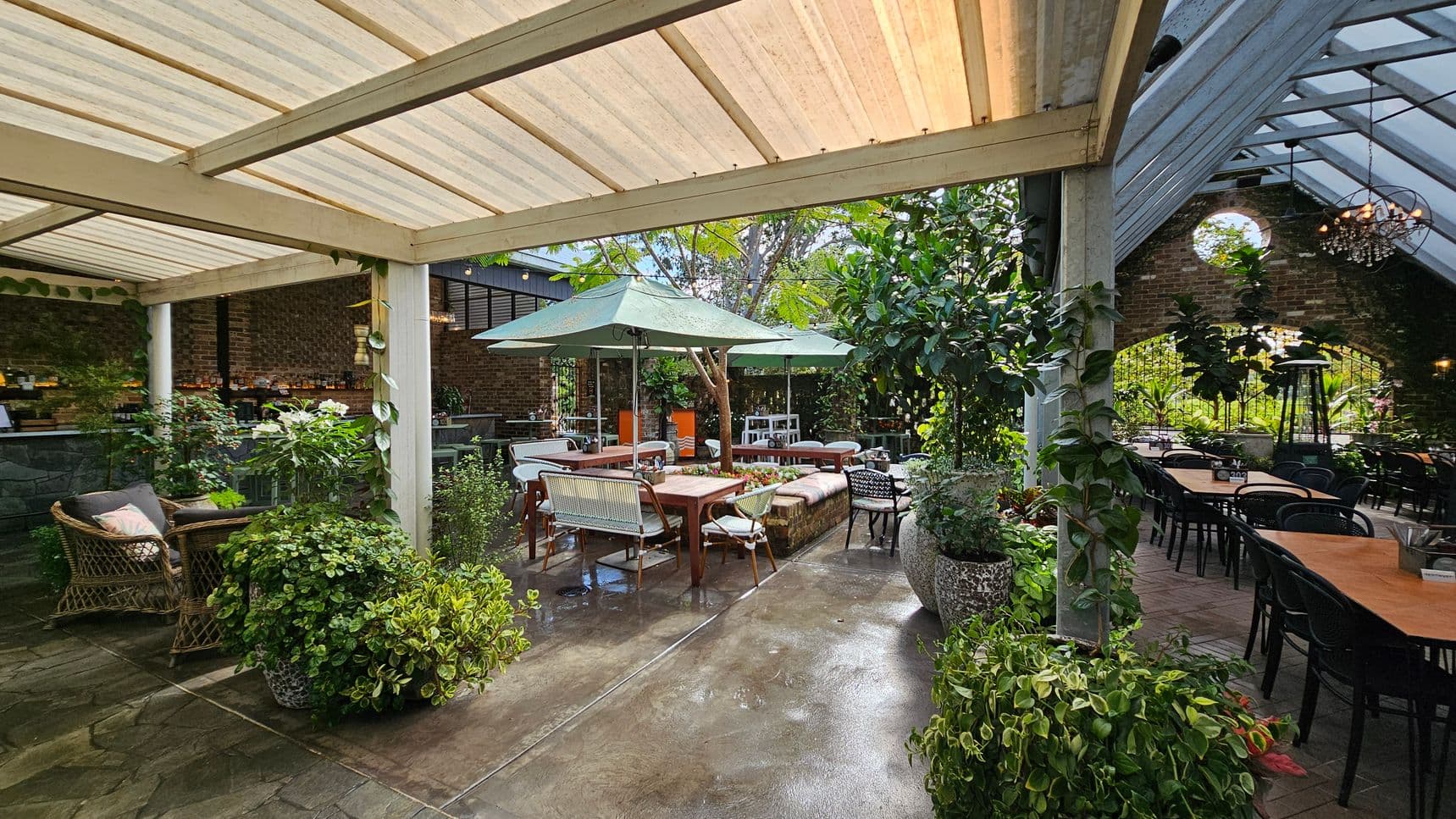 A spacious outdoor patio with a mix of seating arrangements. On the left, there are wicker chairs with cushions and lush green plants in pots. In the centre, a wooden table is surrounded by chairs and benches, shaded by large green umbrellas. The right side features a long table with black chairs under a slanted glass roof. The background has a brick wall with a circular window and more greenery. The floor is a mix of stone and brick, and the ceiling is partially covered with translucent panels.