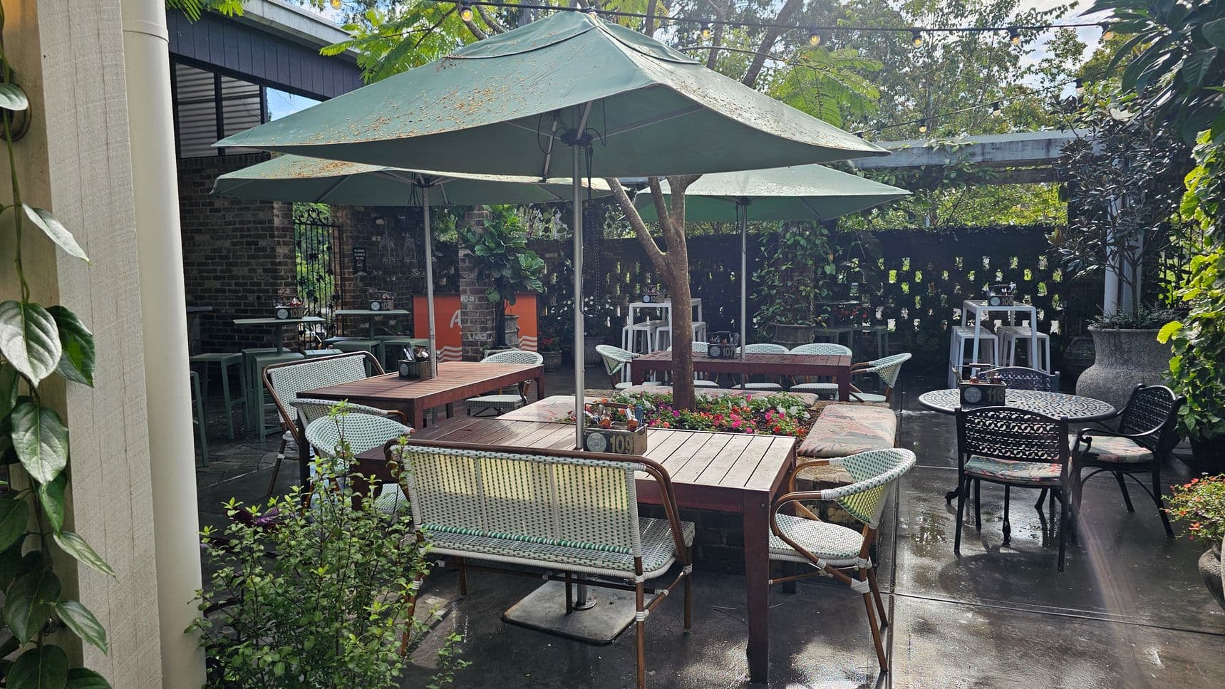 Outdoor seating area with several wooden tables and chairs, some with woven designs. Large green umbrellas provide shade. A variety of plants and flowers are arranged around the space, including a tree in the centre. The ground is paved, and string lights are strung overhead. A brick wall and a wooden structure are in the background, with additional seating and a few stools visible. A large planter is on the right.