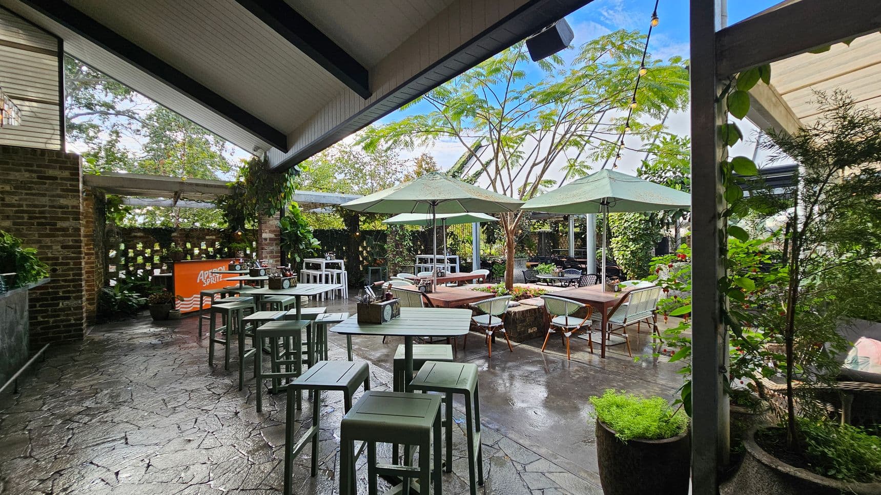 Outdoor seating area with a stone floor and a slanted ceiling. On the left, there's a brick wall with a small bar featuring an "Aperol Spritz" sign. Several high tables with stools are arranged nearby. In the centre, wooden tables with chairs are shaded by large green umbrellas. Lush greenery and potted plants surround the space, with trees visible in the background. String lights are hung across the area, adding a decorative touch.