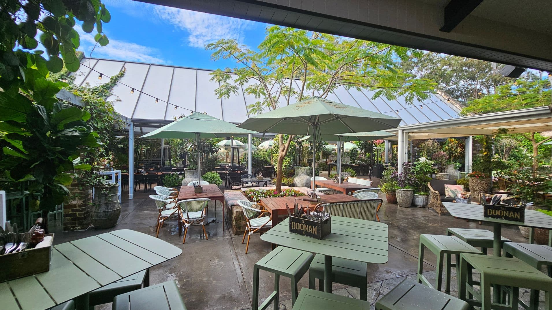 Outdoor seating area with green tables and chairs, surrounded by lush plants. Large umbrellas provide shade over wooden tables. A tree is centrally located, with string lights overhead. The background features a glass-roofed structure and more greenery. The floor is paved, and there are decorative planters throughout.