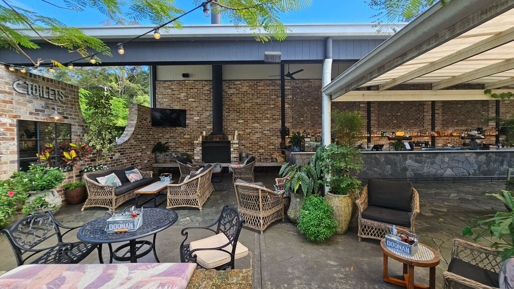 Outdoor seating area with wicker furniture and a brick wall backdrop. On the left, a sign for toilets is visible on the brick wall, surrounded by potted plants. In the centre, a fireplace is flanked by a TV and more seating. On the right, a bar with a stone facade is under a covered area. String lights hang above, and the floor is paved with stone tiles.