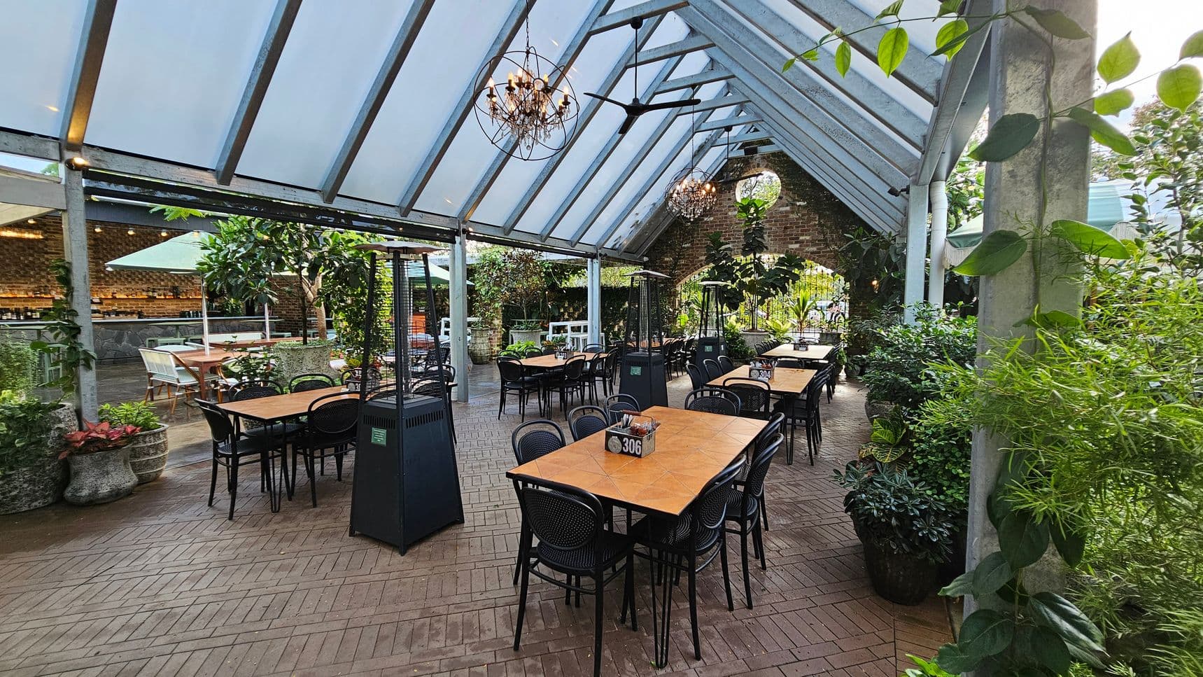 A spacious outdoor dining area with a glass roof and brick flooring. In the centre, there are several wooden tables with black chairs, each marked with a number. On the left, a bar area with a brick wall and a large umbrella. On the right, lush greenery and potted plants line the space. Hanging chandeliers and ceiling fans are suspended from the roof. The atmosphere is open and inviting, with a mix of natural and industrial elements.