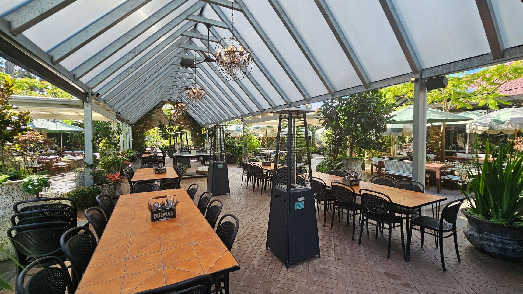 A spacious outdoor dining area with a glass roof supported by metal beams. Long wooden tables with black chairs are arranged in rows. Two tall outdoor heaters are positioned along the centre aisle. The floor is paved with bricks in a herringbone pattern. Lush greenery and potted plants surround the space, with additional seating visible in the background under green umbrellas. Chandeliers hang from the ceiling, adding a decorative touch.