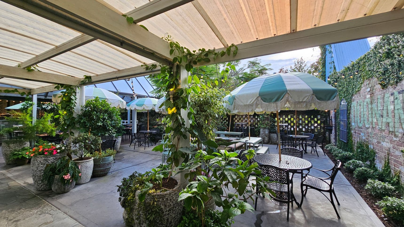 Outdoor patio with a covered pergola, featuring climbing plants on the beams. Several large potted plants with vibrant flowers are scattered around. Black metal tables and chairs are arranged under green and white striped umbrellas. A brick wall in the background is partially covered with ivy, and string lights are hung above. The floor is paved with stone tiles.