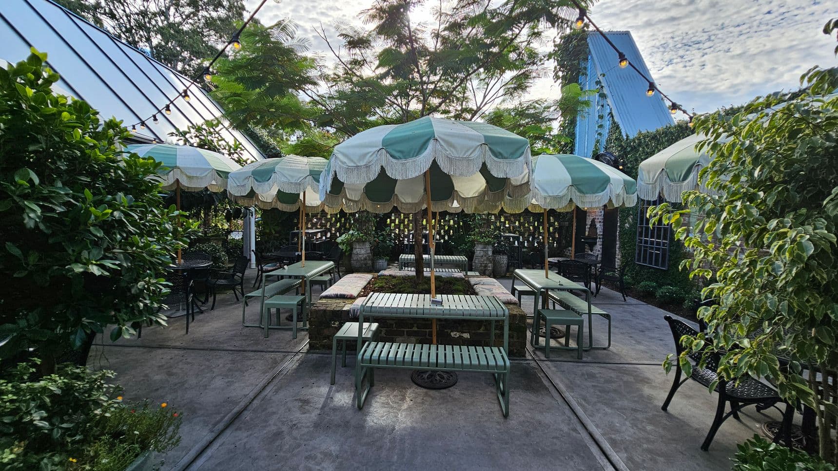 Outdoor seating area with green and white striped umbrellas in the centre, surrounded by green metal benches and tables. The ground is concrete, and there are string lights overhead. Lush greenery and plants border the area on the left and right, with a blue-roofed structure visible in the background. The setting has a garden-like atmosphere with a mix of natural and architectural elements.