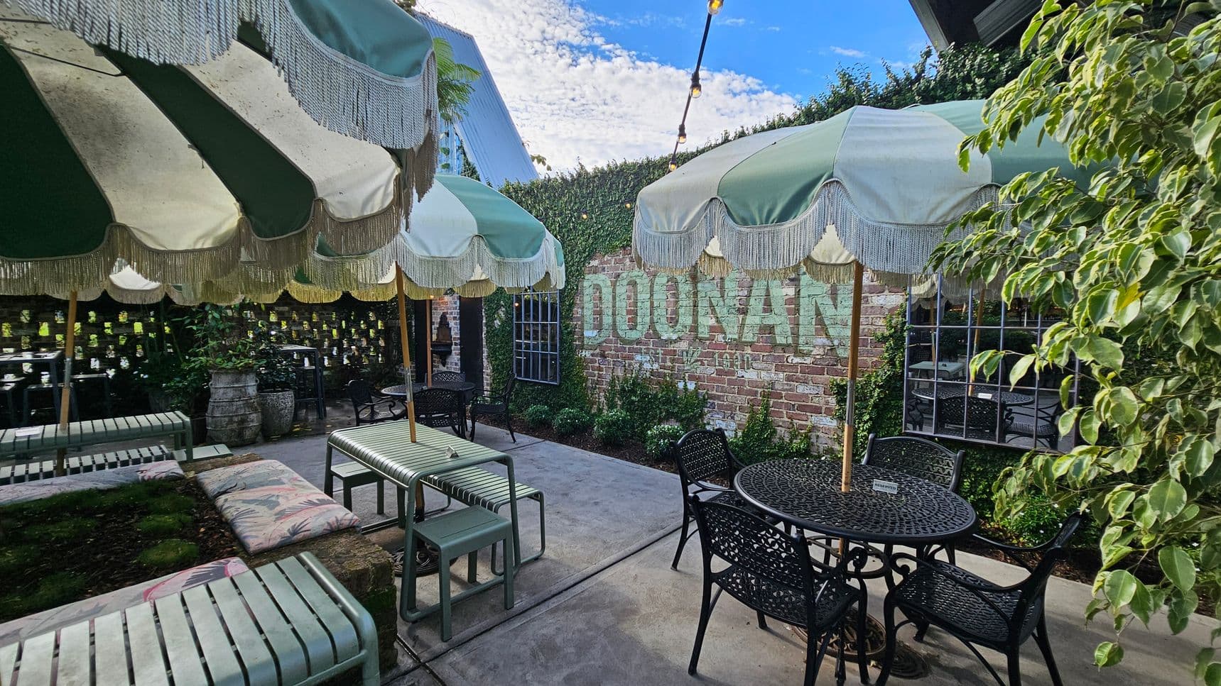 A courtyard with green and white striped umbrellas featuring fringe, providing shade over metal tables and chairs. On the left, a bench with a floral cushion is visible. The background has a brick wall with the word "DOONAN" painted on it, partially covered by ivy. A window with a grid pattern is set into the wall. The right side has a leafy plant, and string lights are strung across the area. The floor is concrete.