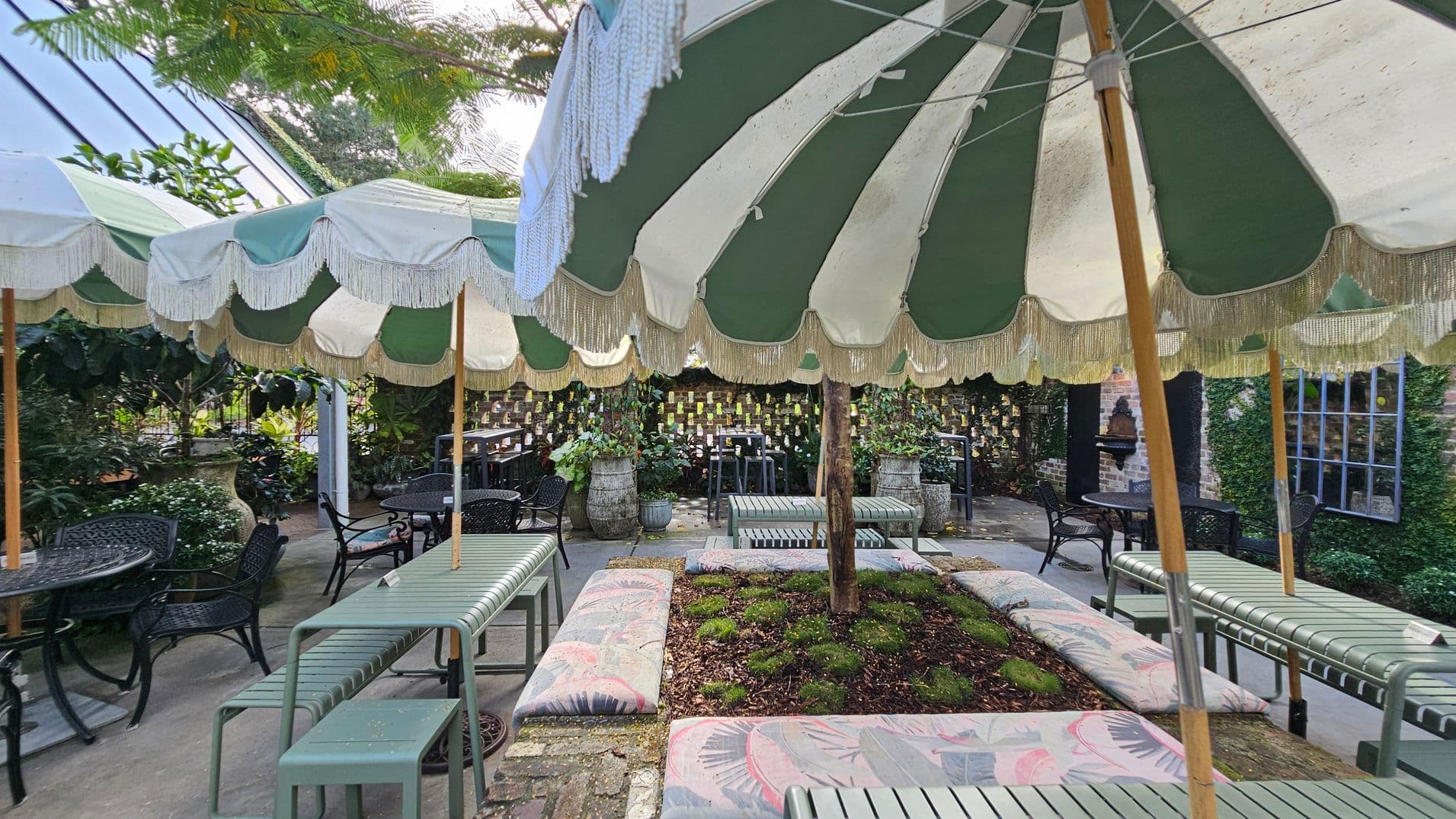A garden setting with green and white striped parasols featuring fringe, providing shade over seating areas. The seating includes metal benches and cushioned seats around a small garden bed with plants. In the background, there are potted plants and a brick wall adorned with small hanging items. Tables and chairs are scattered throughout, with greenery and foliage visible around the perimeter. The floor is paved with stone tiles.