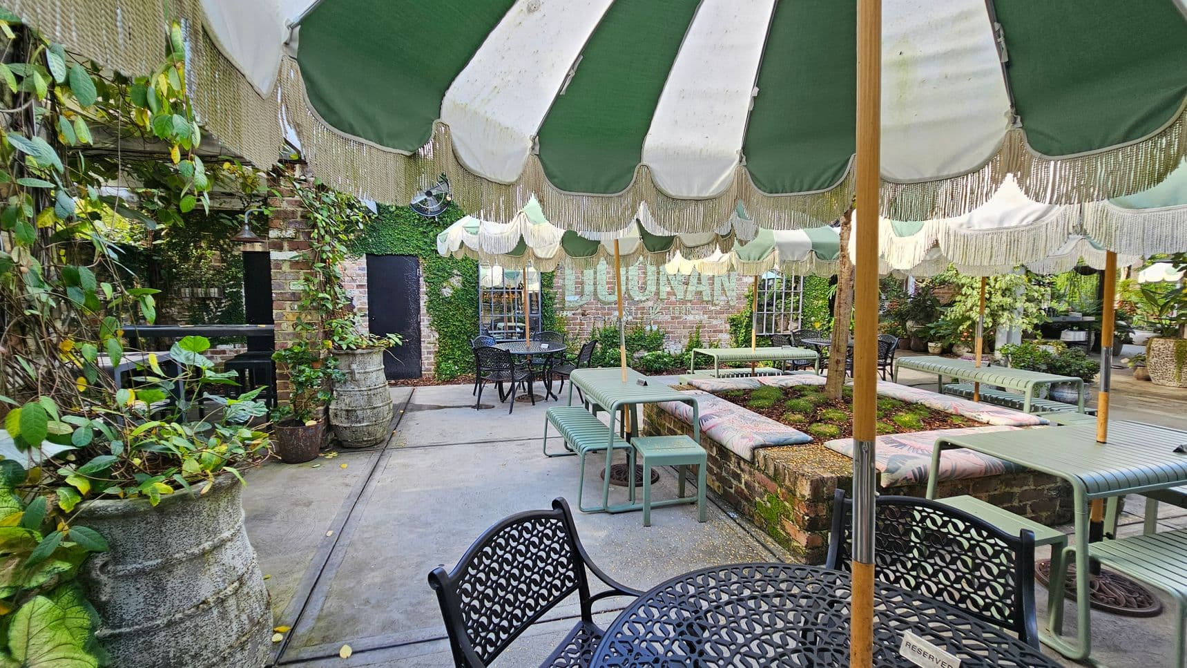 Outdoor seating area with green and white striped umbrellas providing shade. On the left, there are large planters with climbing plants against a brick wall. In the centre, metal tables and benches are arranged around a raised garden bed. The background features a brick wall with ivy and a sign partially visible. The floor is concrete, and the area is surrounded by lush greenery.