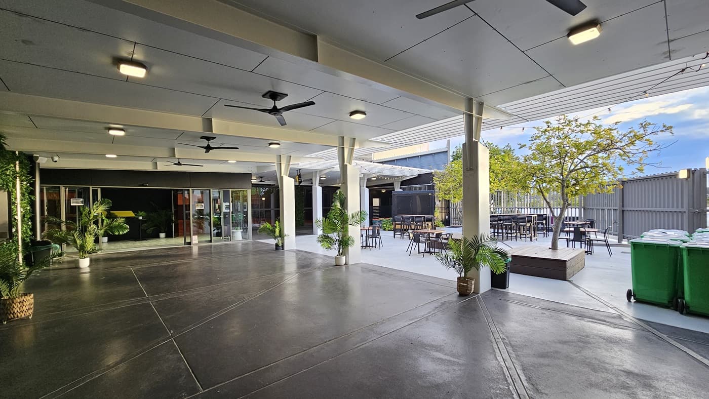 On the left, there is a glass entrance with potted plants and a black wall. On the right, there is an outdoor seating area with tables, chairs, and a tree in a planter box. In the middle, there is an open space with a concrete floor and several ceiling fans. The background features a grey fence and additional seating. The ceiling is white with recessed lights and fans. Green bins are placed near the fence on the right.