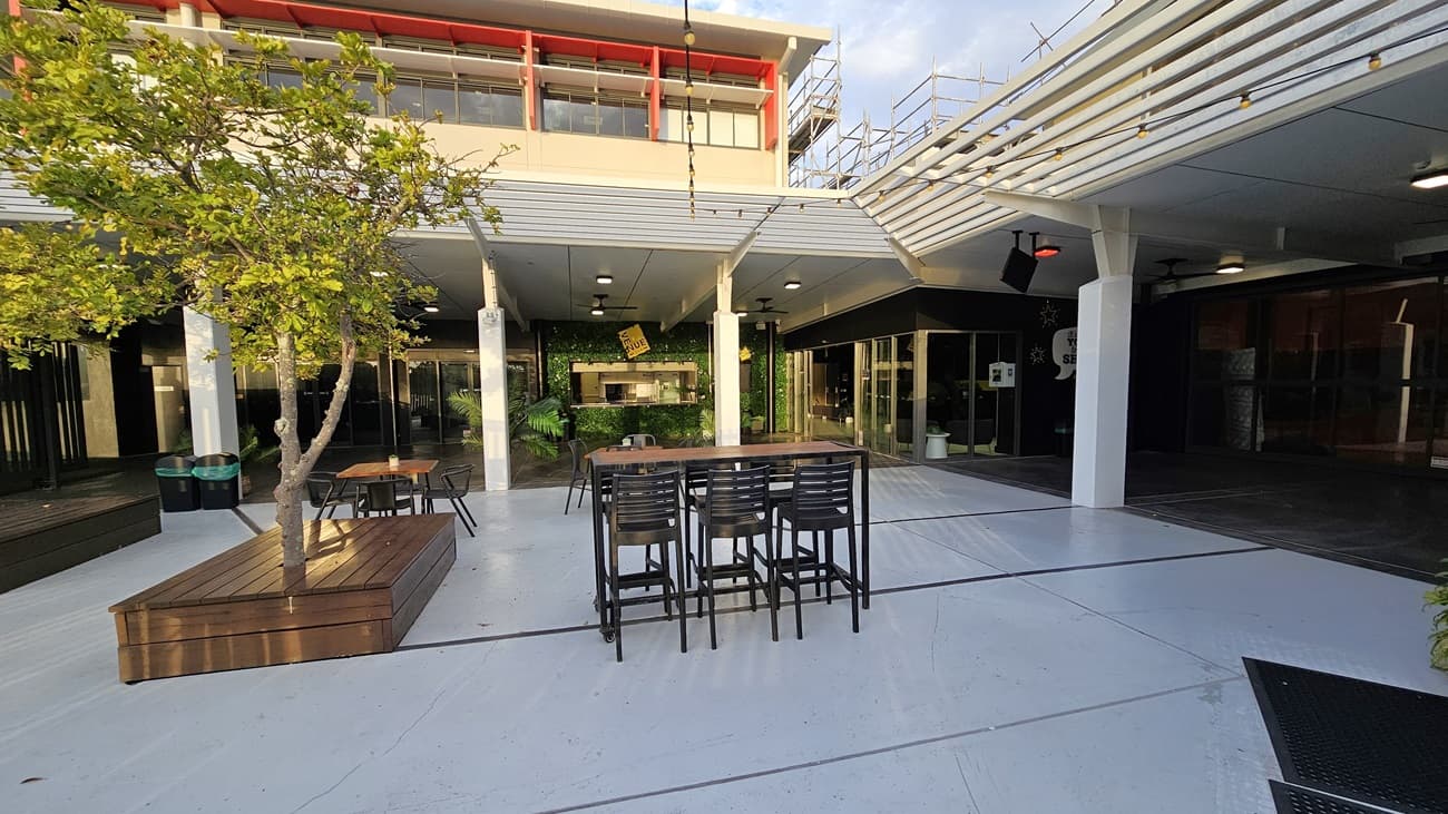 On the left, there is a wooden bench surrounding a small tree and two green bins. On the right, there is a covered area with black walls and glass doors. In the middle, there is an outdoor seating area with a tall table and several chairs. The background features a building with large windows and a red trim on the upper floor. The floor is light grey concrete, and the ceiling of the covered area is white with recessed lights.