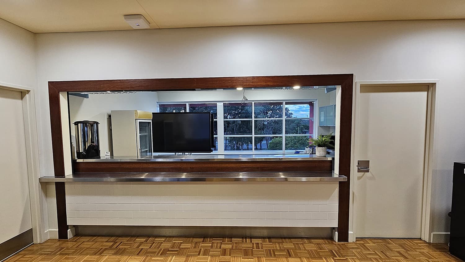 On the left, there is a white door with a silver handle. On the right, there is another white door with a silver handle and a black cabinet next to it. In the middle, there is a large rectangular mirror with a wooden frame above a long stainless steel counter. Behind the mirror, there is a television mounted on the wall and a potted plant on a shelf. The floor is covered in wooden parquet, and the ceiling is beige with recessed lighting.