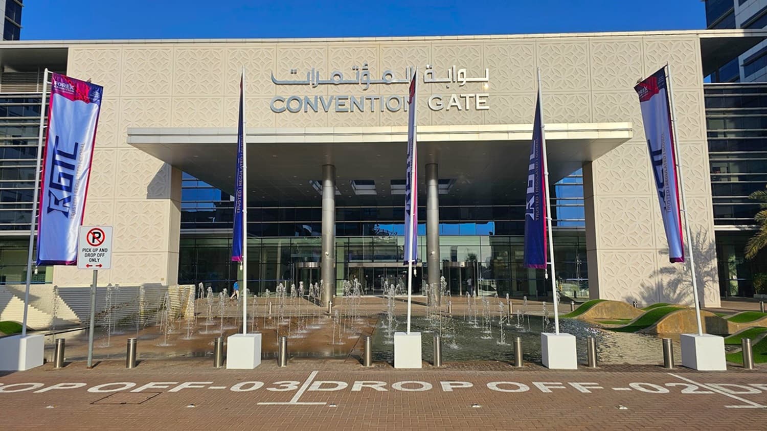 A modern building with a glass facade on the right, featuring large windows and a sleek design. On the left, there are palm trees lining a paved walkway. The building has a decorative patterned wall and a large overhang supported by metal columns. The ground is covered with interlocking bricks, and there are bollards along the path. Shadows of the palm trees are cast on the ground.